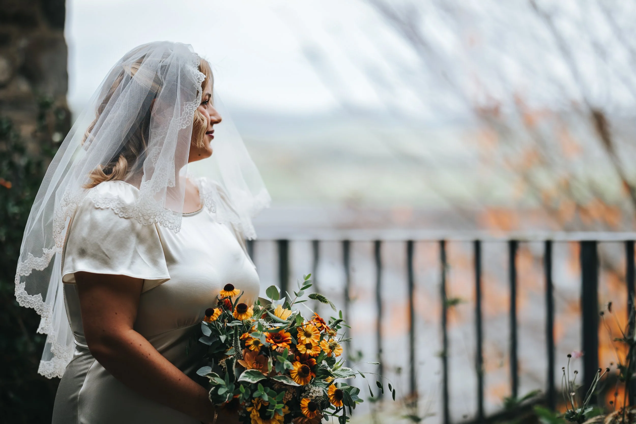 A woman wearing a white wedding dress and veil holding a bouquet of yellow and orange flowers, standing near an outdoor railing with out-of-focus trees in the background.