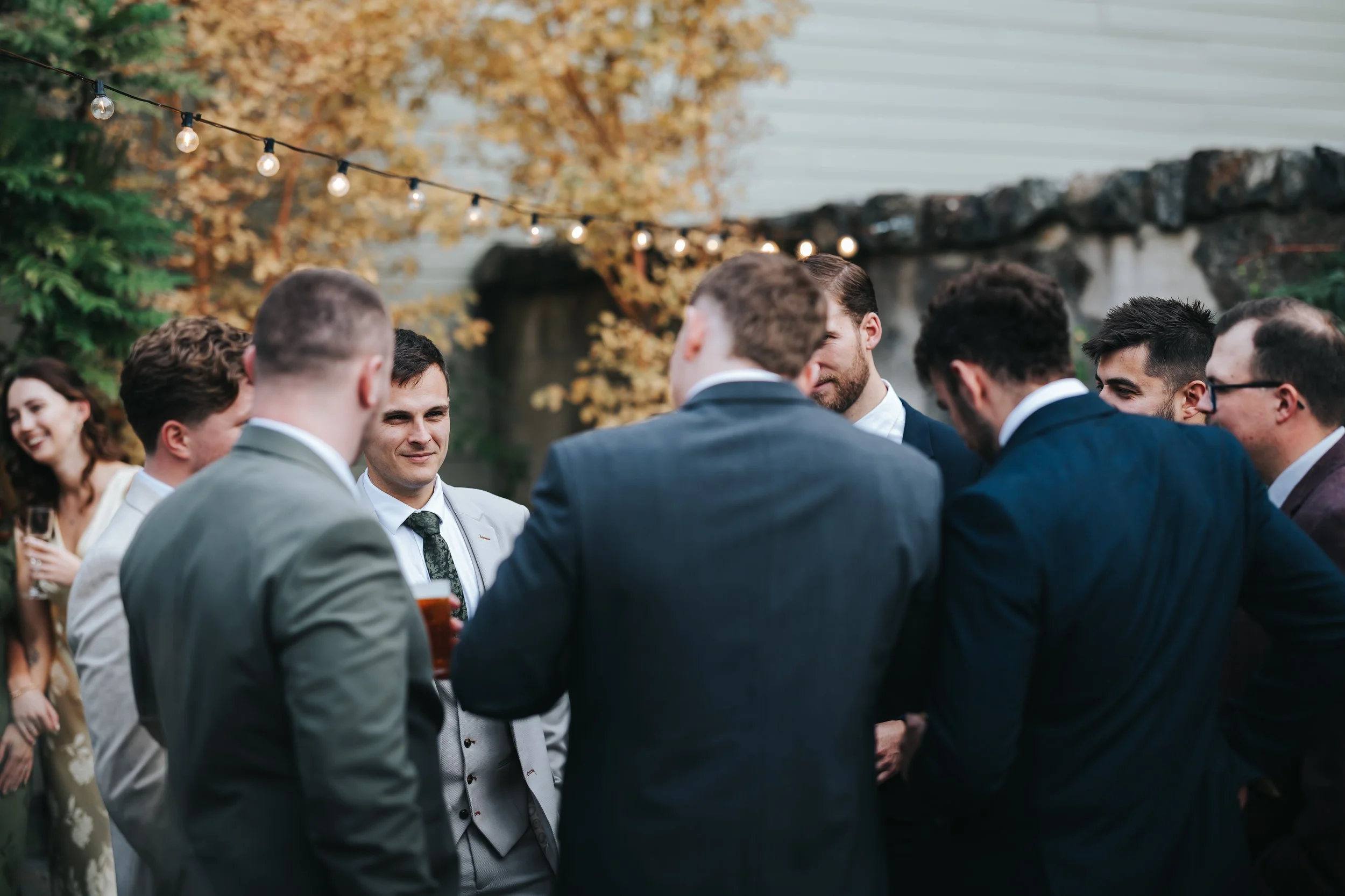Group of men and women in suits and dresses socializing at an outdoor event during the evening, with string lights overhead and autumnal foliage in the background.