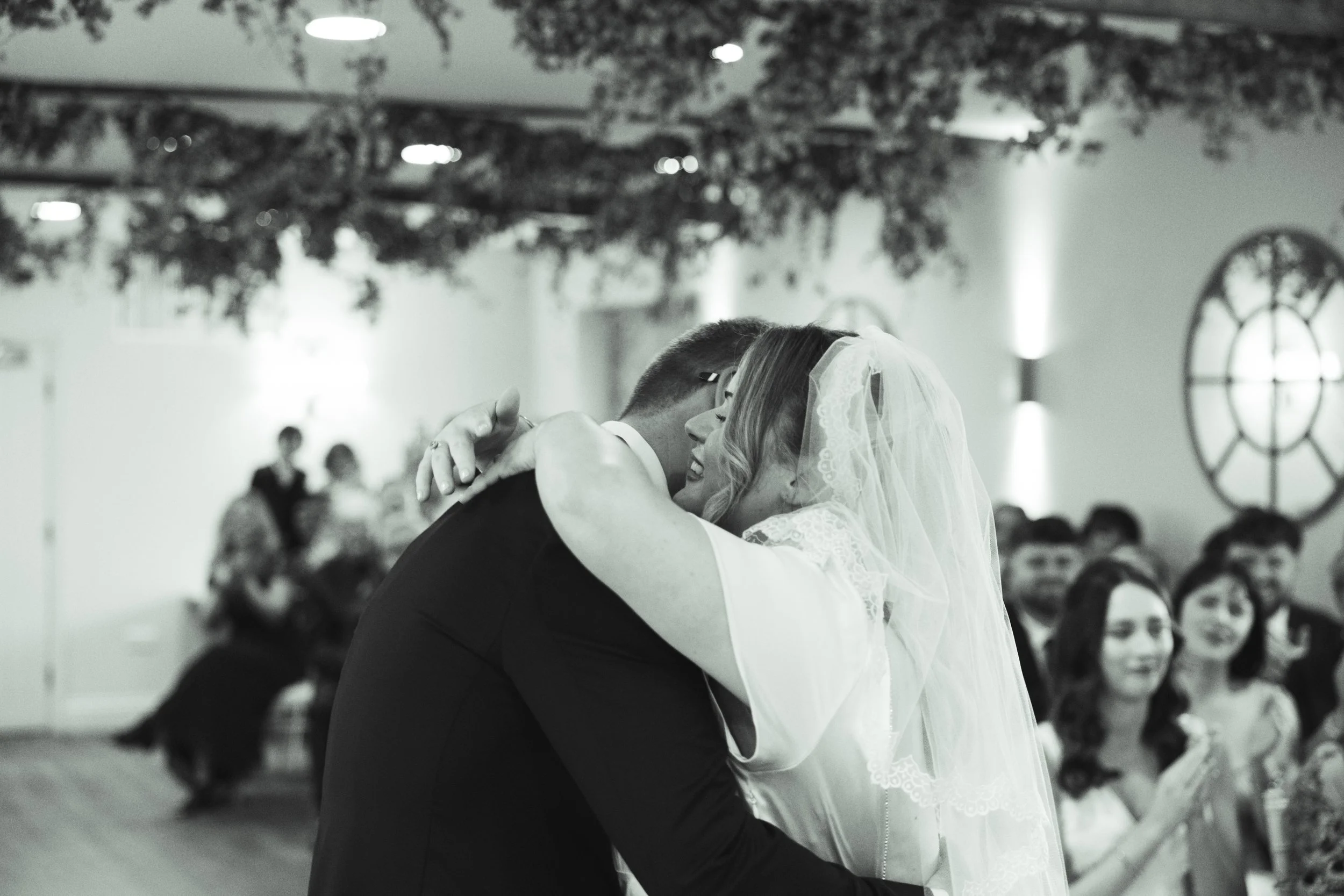 A bride and groom share a kiss during their wedding ceremony indoors, with guests seated in the background watching and smiling.