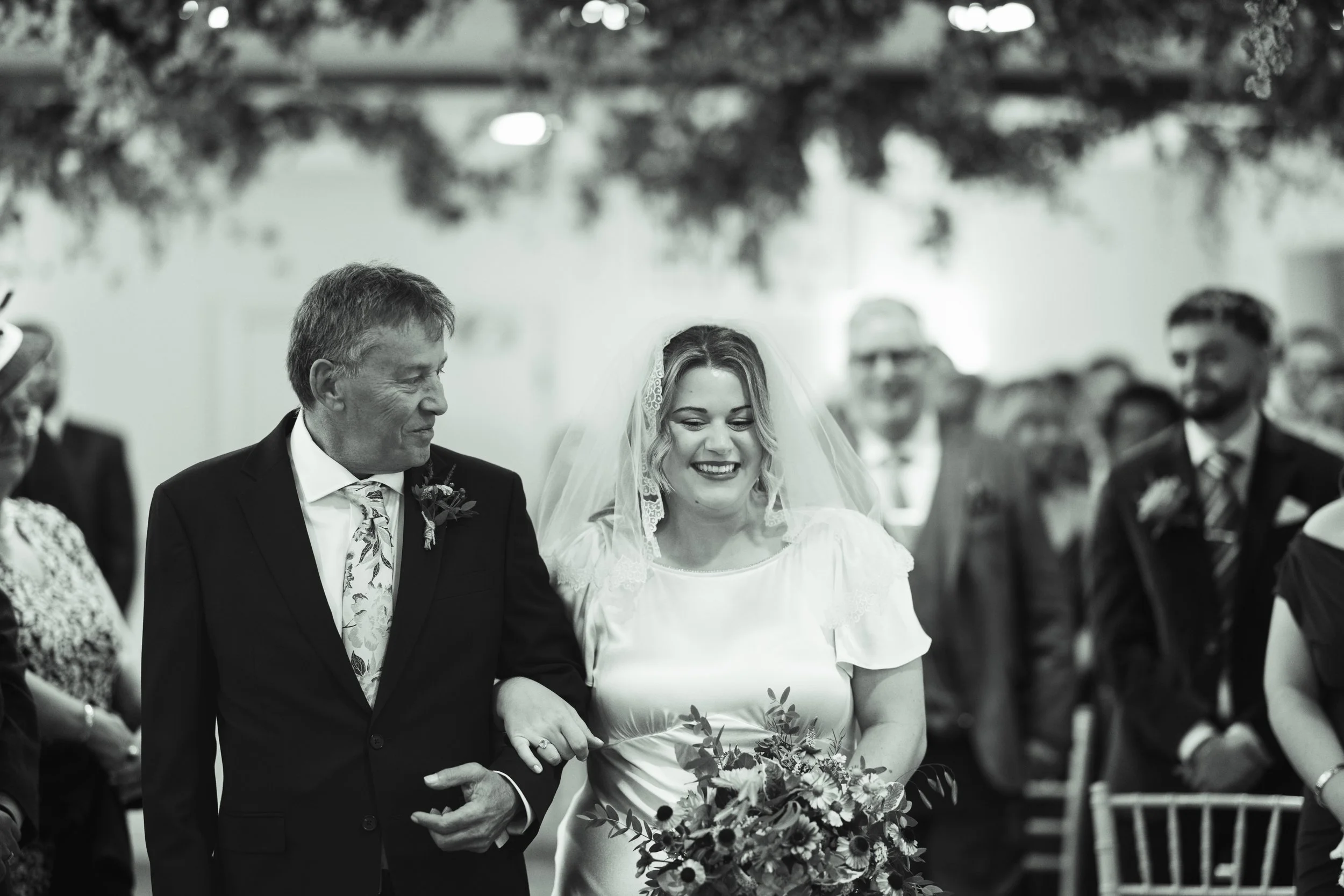 A bride exiting a ceremony with her arm linked with an older man, possibly her father, in a room filled with seated guests. The bride wears a wedding dress and veil, holding a bouquet, and is smiling. The guests are dressed in formal attire.