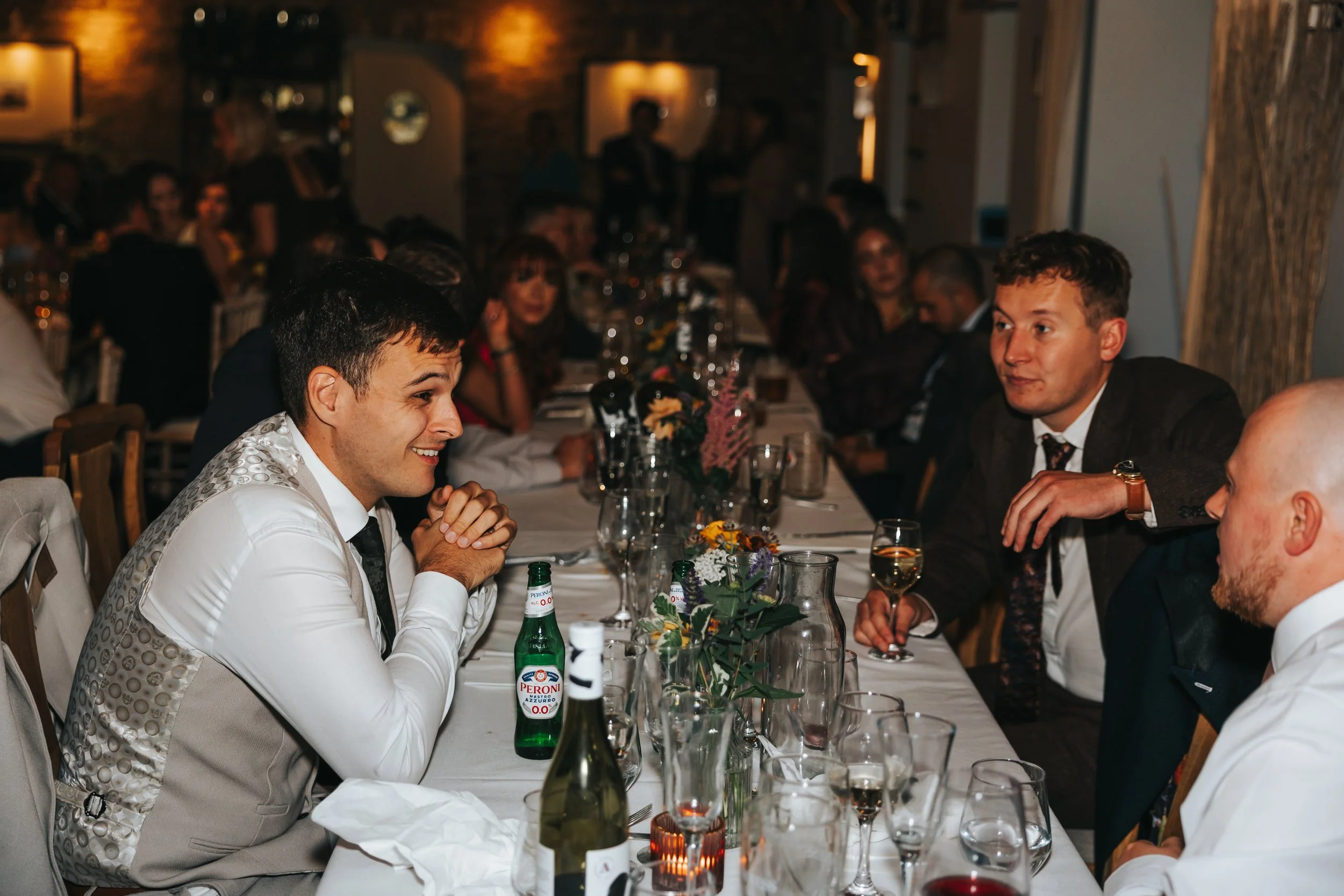 People sitting at a dinner table, engaged in conversation, during a social event or celebration in a warmly lit restaurant or banquet hall.