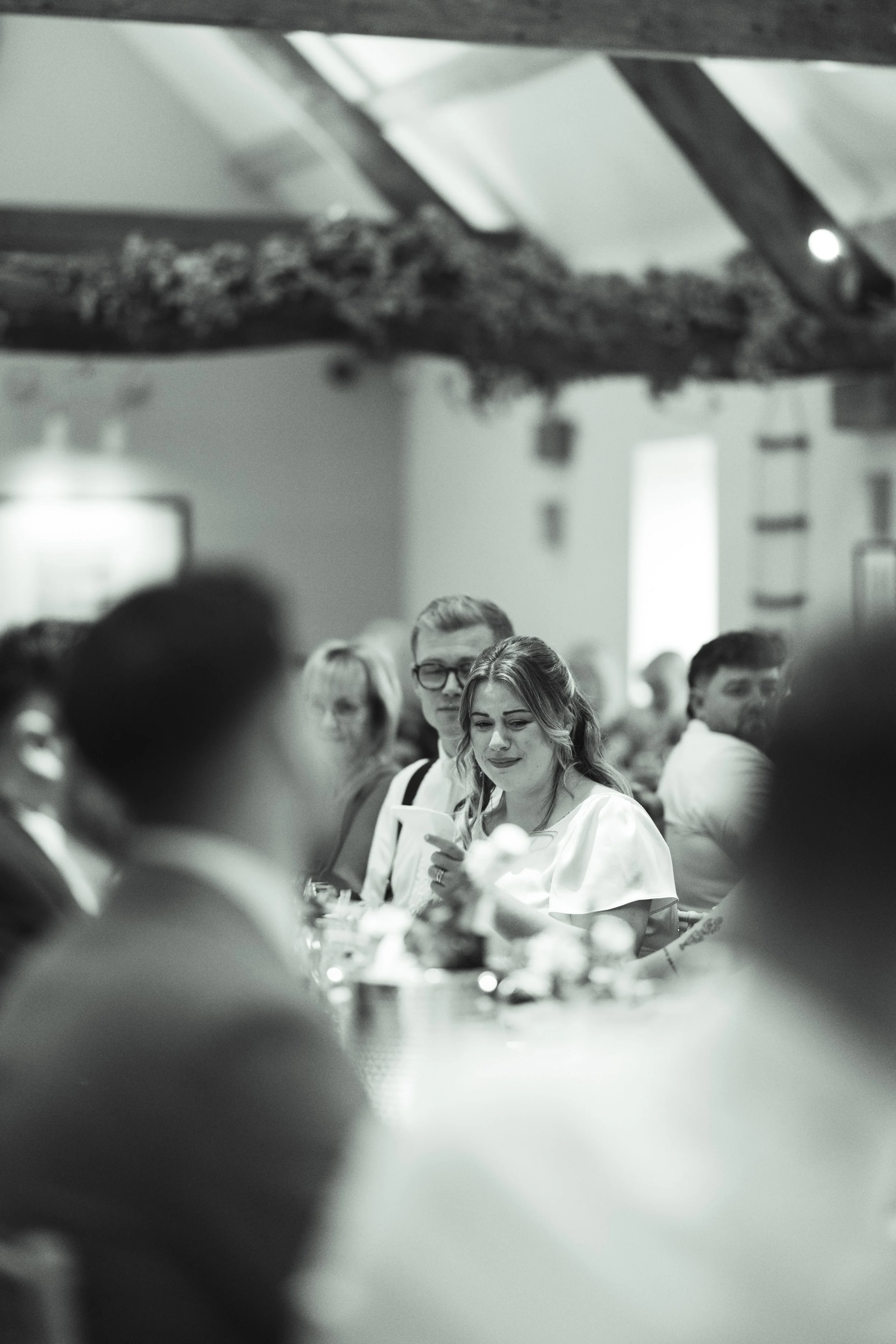 People gathered at a social event or celebration, with one woman in focus using her phone, in a dimly lit indoor setting in black and white.