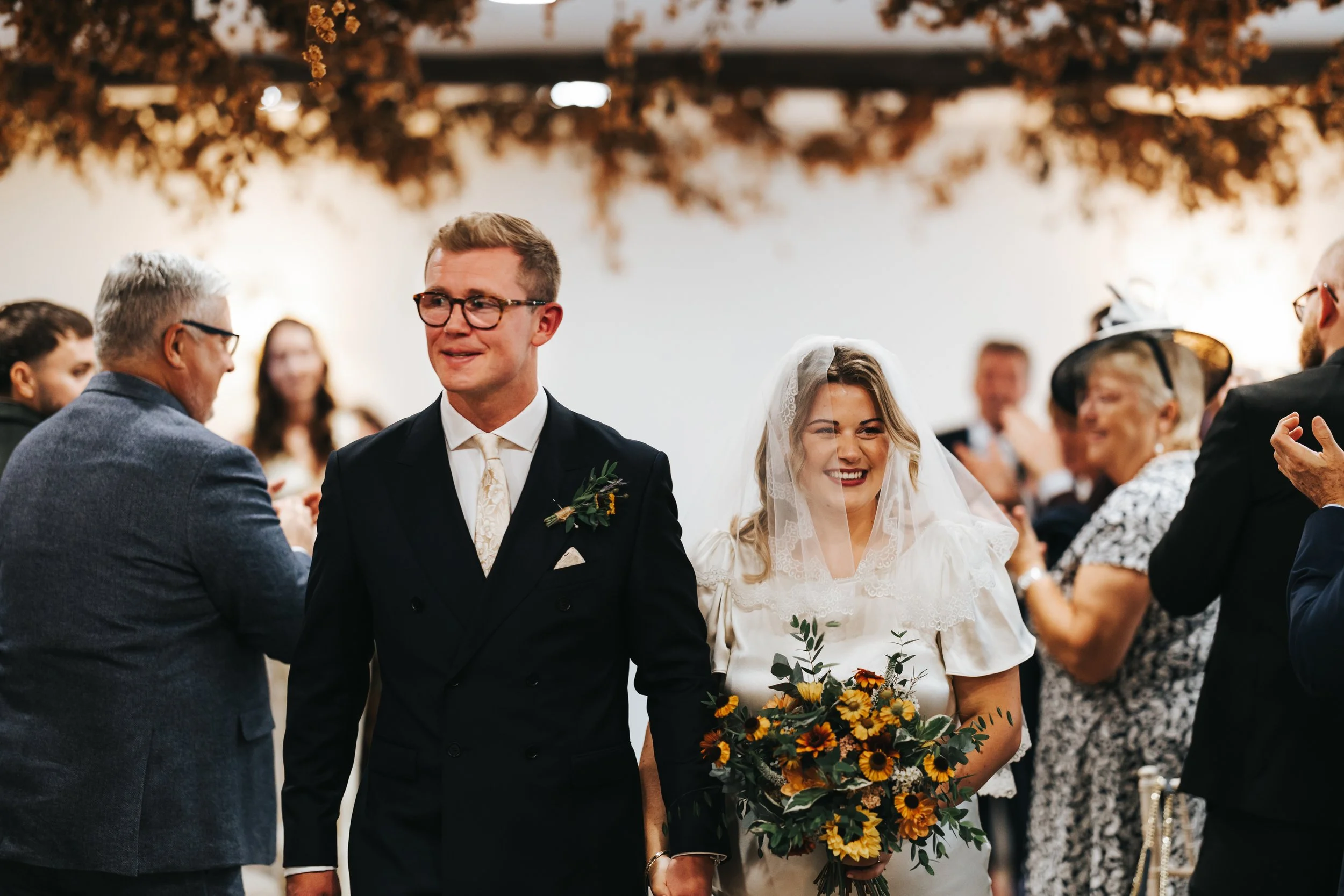 A bride and groom walking down the aisle at their wedding, smiling. The bride is holding a large bouquet of sunflowers and greenery. Guests are clapping and celebrating around them.