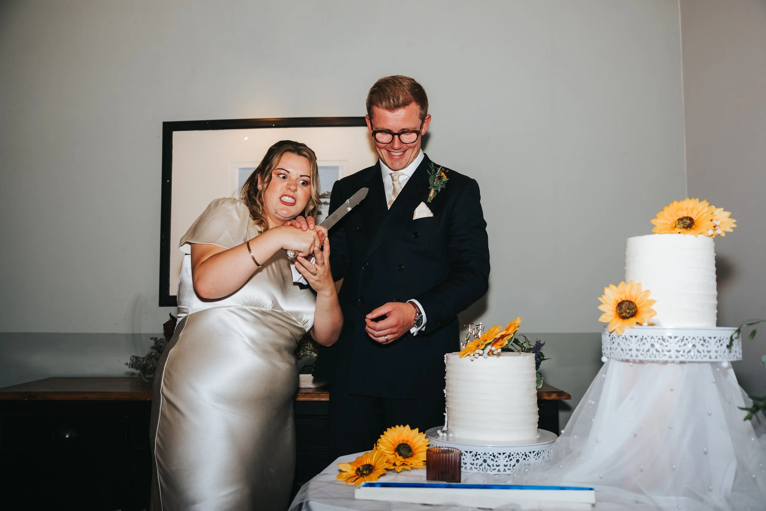 A woman in a champagne dress and a man in a black suit with glasses cut a wedding cake with sunflowers, while the woman looks fearful.