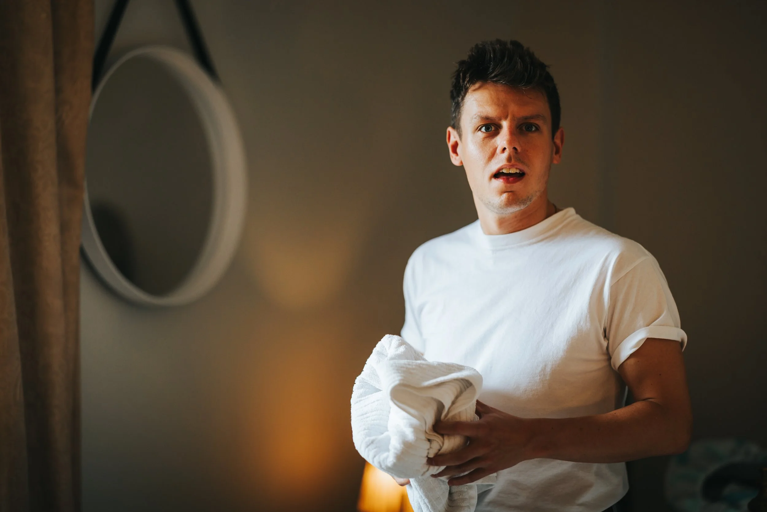 A surprised young man with dark hair wearing a white t-shirt holding a folded towel in a dimly lit room.
