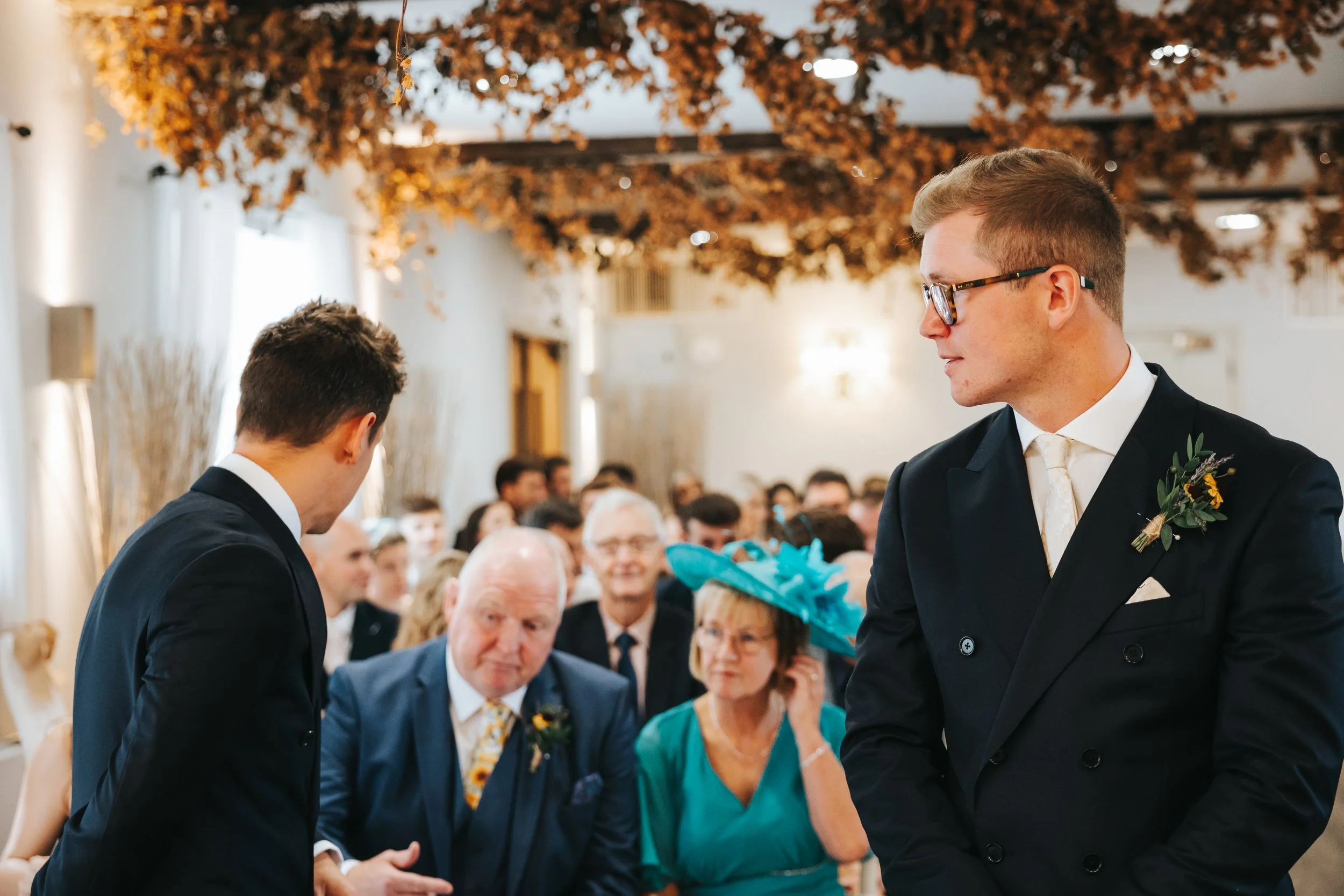 A wedding ceremony with two grooms in suits, one wearing glasses, engaging in the vows. The bride and groom are in focus, with an elderly man and woman, and other guests watching in the background. The venue has decorative brown hanging leaves and wa