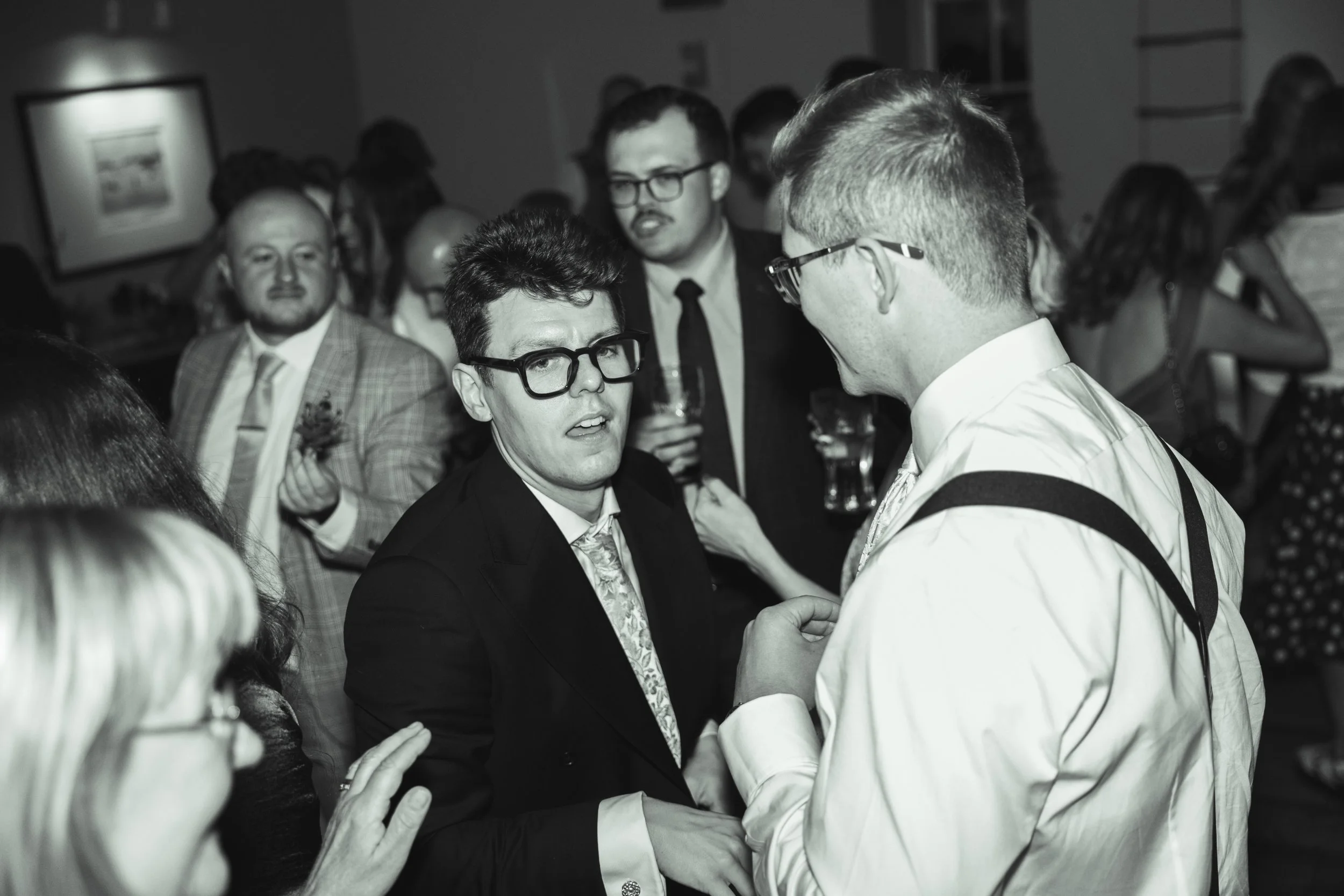 A group of people at a formal event, with two men in the foreground shaking hands and engaging in conversation, one wearing glasses and a suit, the other with glasses, a white shirt, and suspenders. Others in the background hold drinks and converse.
