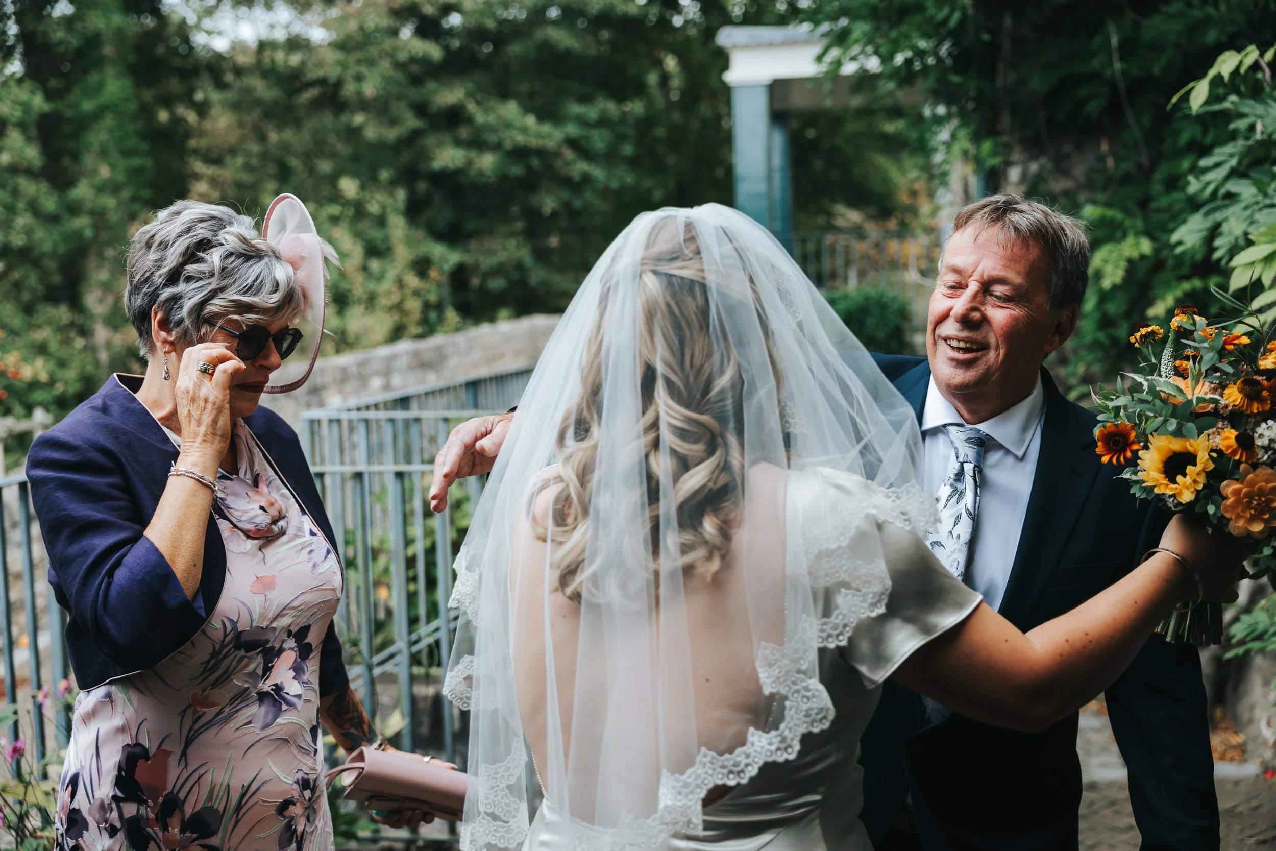 A bride with blonde hair in a white dress and veil is holding a bouquet of sunflowers and talking to an elderly woman with glasses, short gray hair, a hat, and a floral dress. A man in a suit is smiling and touching the bride's shoulder.