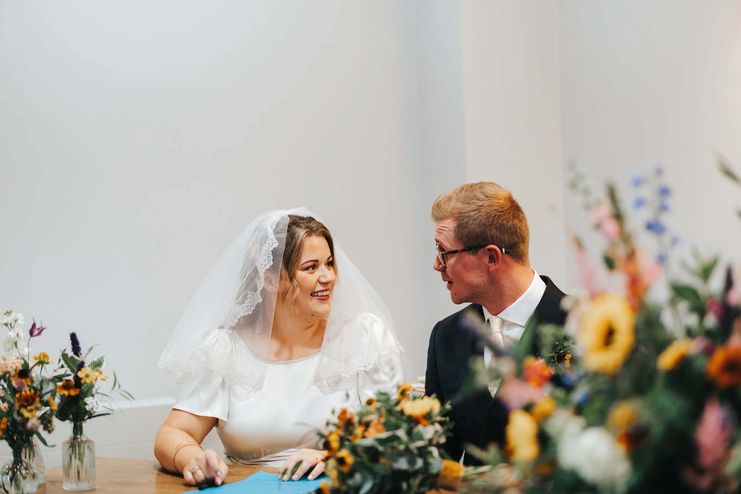 A bride and groom sitting at a table during their wedding ceremony, looking at each other and smiling, with colorful flowers in vases in front of them.