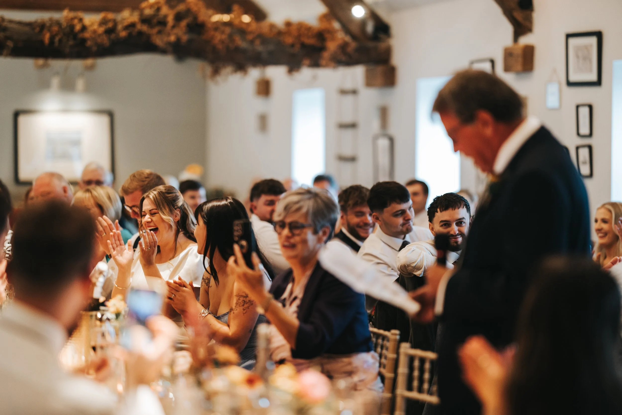 A man giving a speech at a wedding reception surrounded by guests who are smiling, laughing, and taking photos, in a warmly decorated indoor setting.