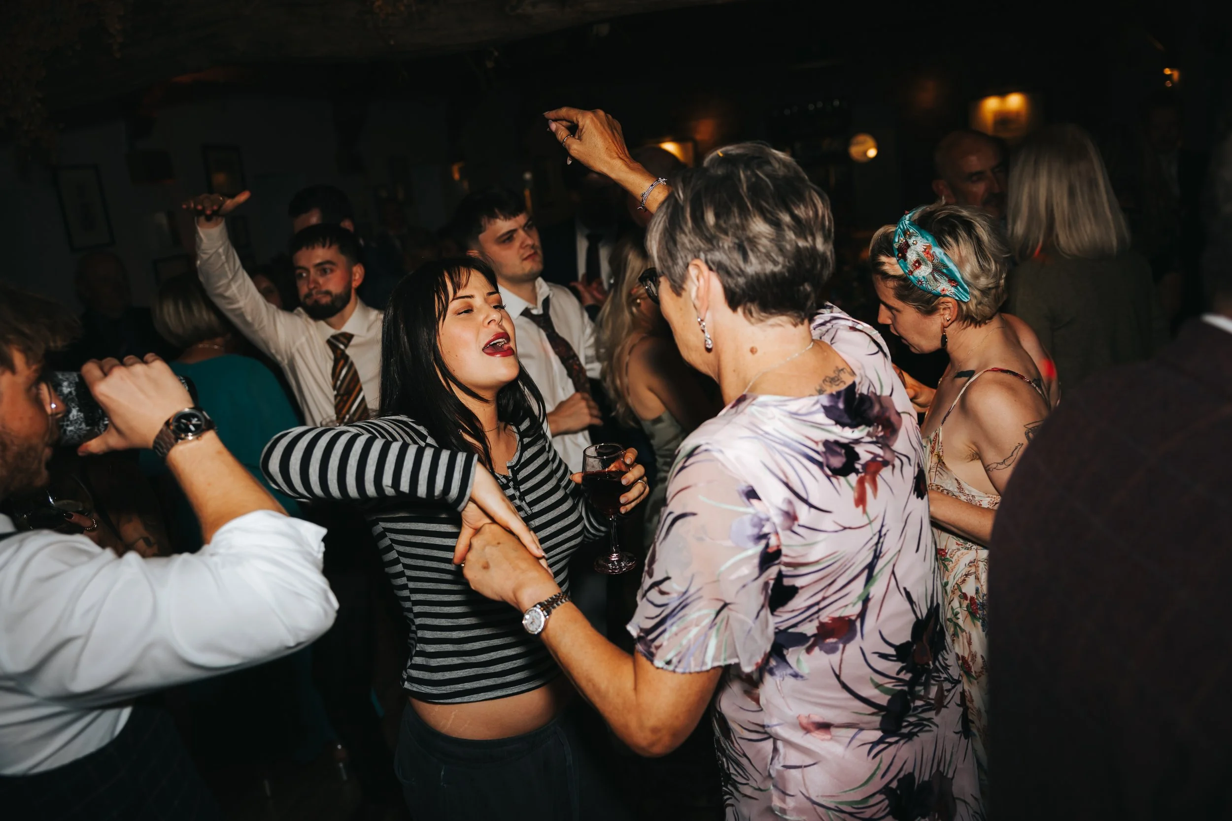 Group of people dancing and socializing at a lively party or nightclub, some holding drinks, in a dimly lit indoor setting.