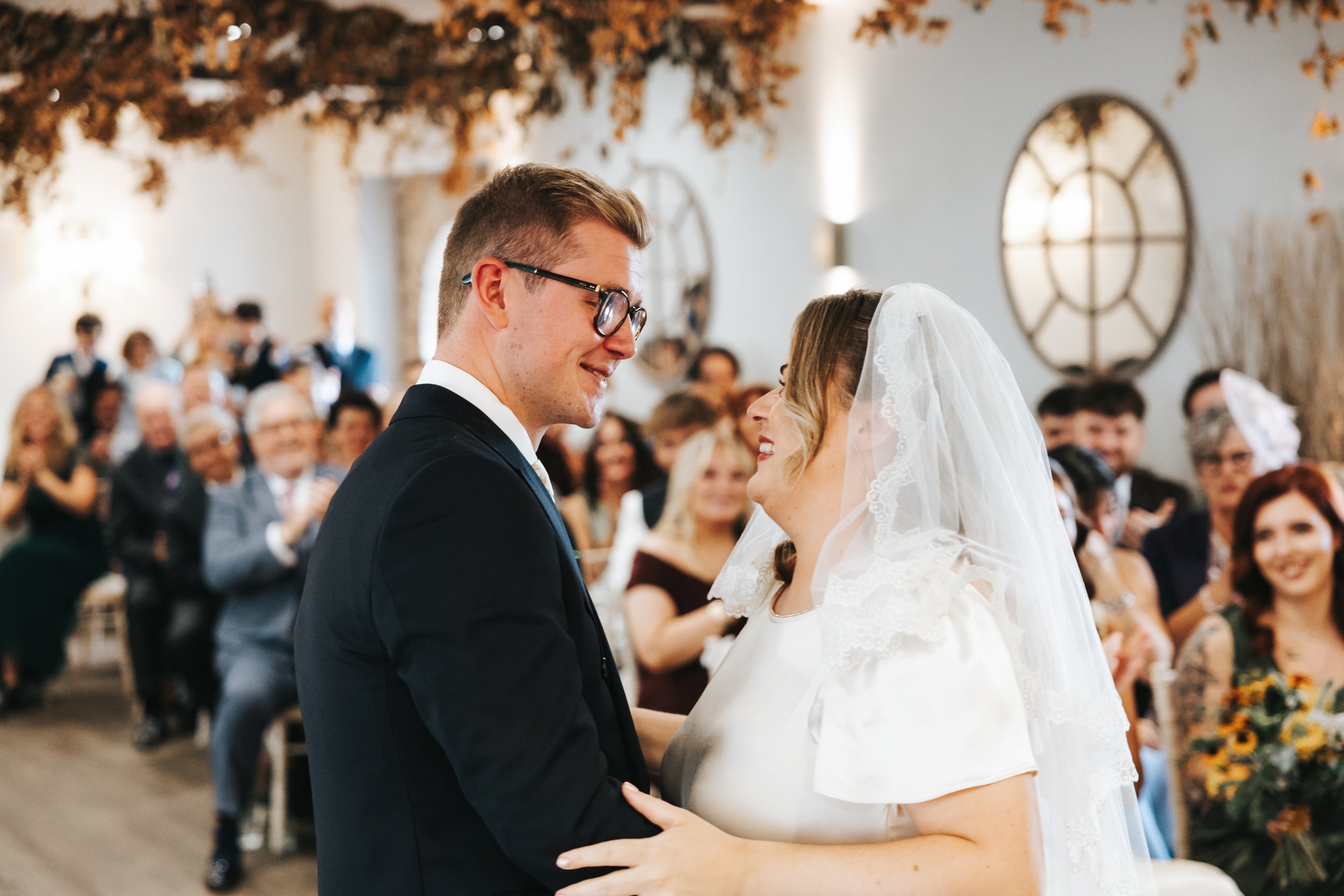 A bride and groom smiling at each other during their wedding ceremony, surrounded by seated guests in an indoor venue decorated with fall leaves and large circular wall ornaments.