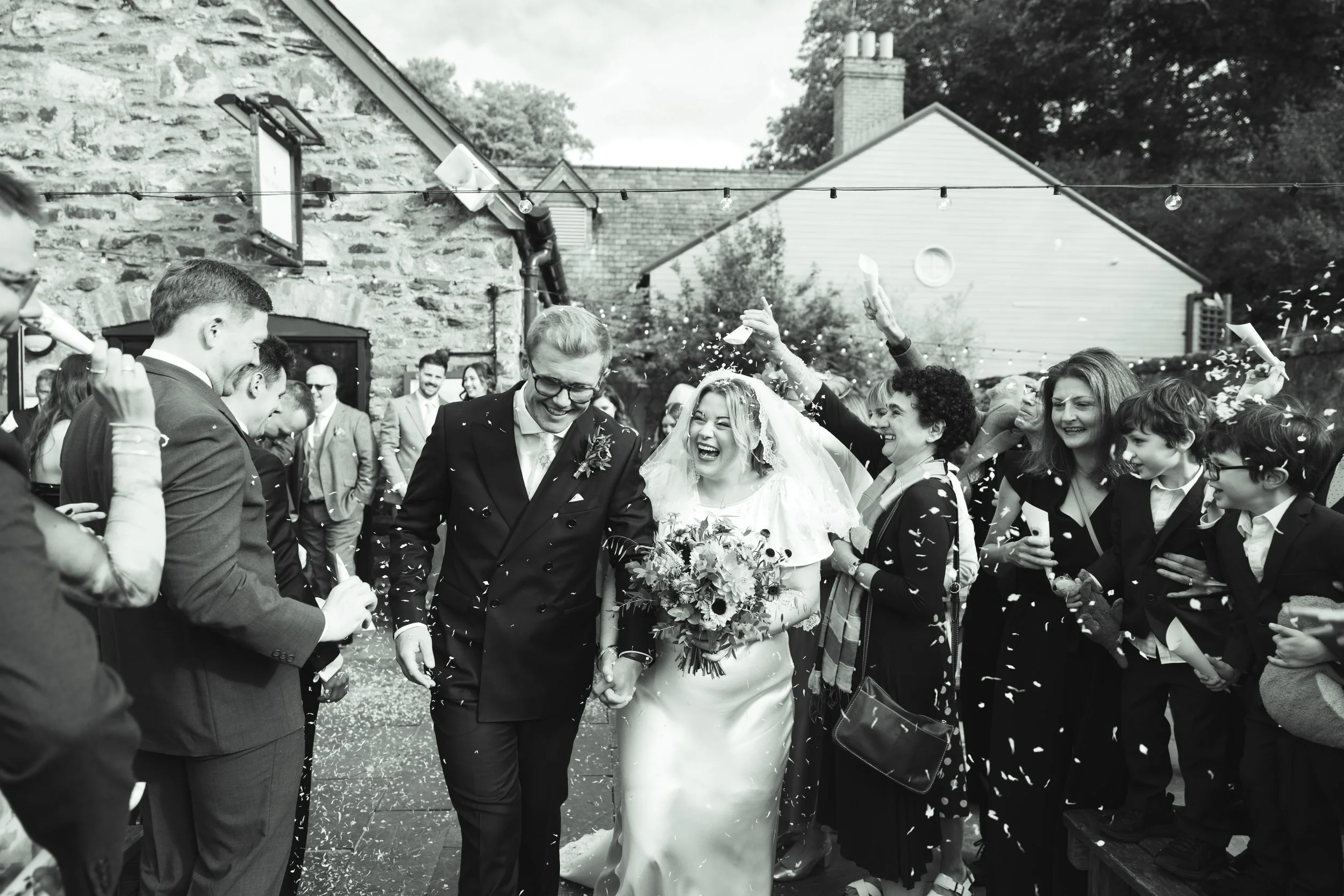 Black and white photo of a wedding celebration, with a bride and groom walking hand in hand through a crowd of people throwing confetti.