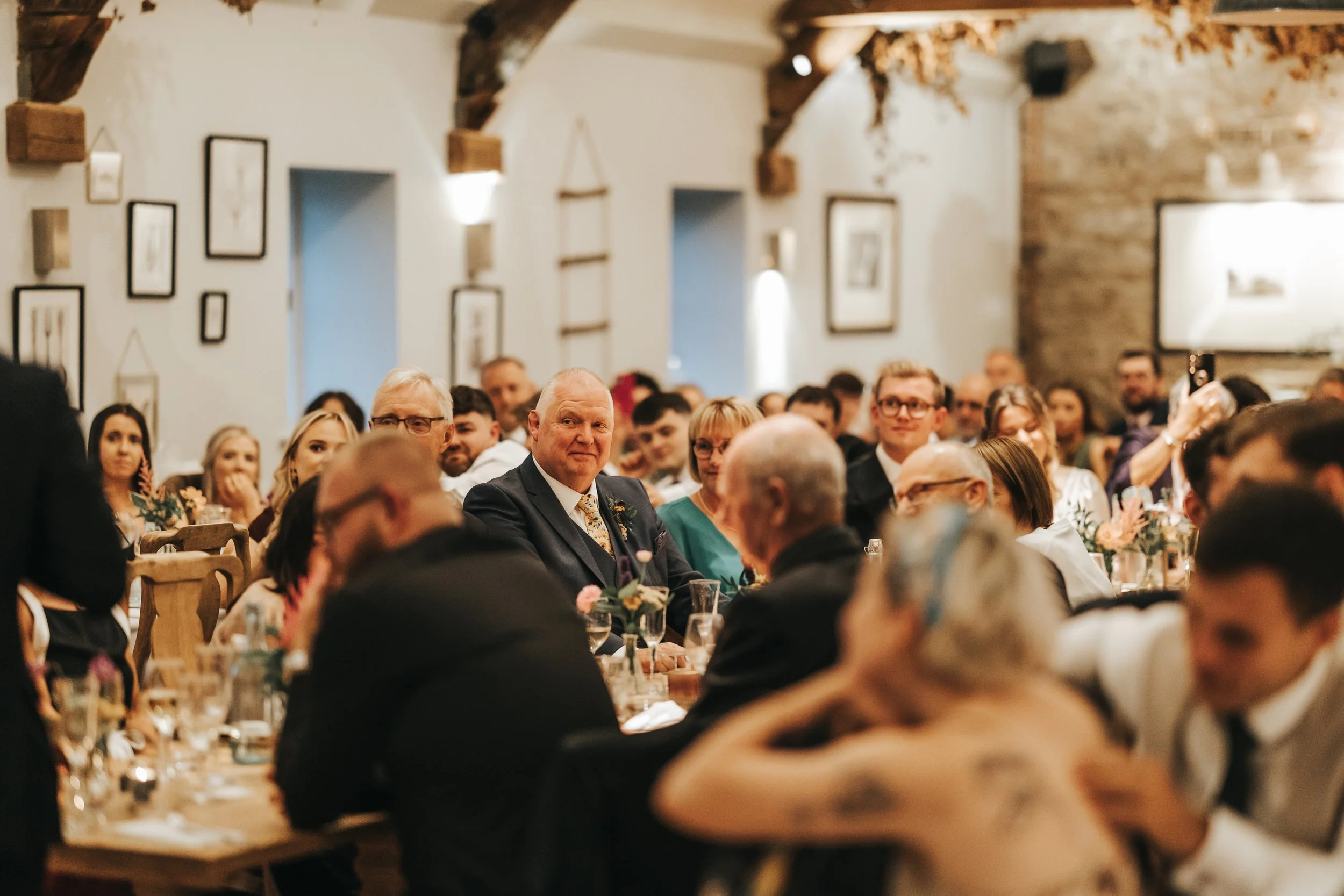 People sitting at tables in a decorated rustic venue, attending a formal event or celebration, with some looking towards the camera.
