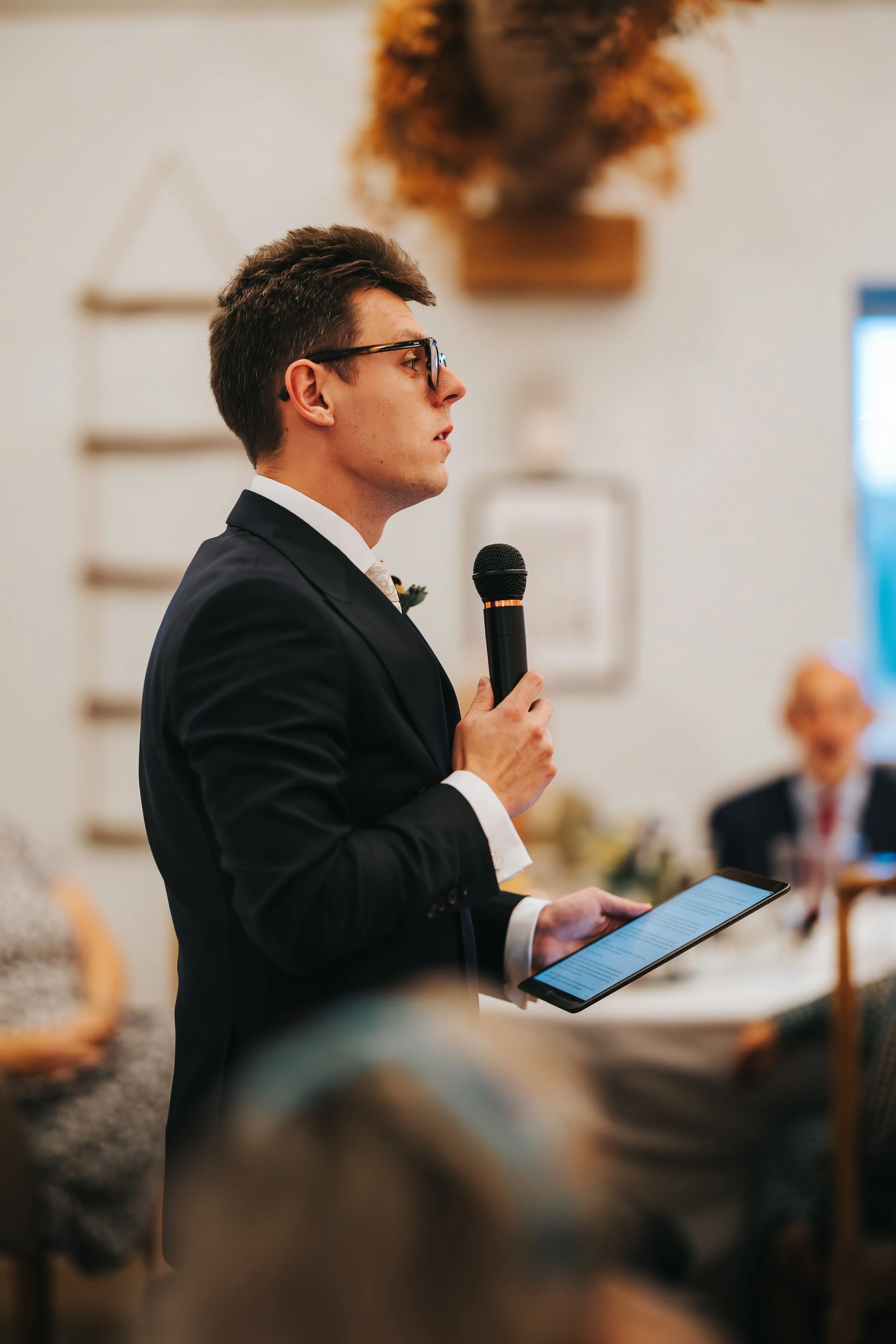 A man in a suit holding a microphone and reading from a tablet, speaking at an indoor event.