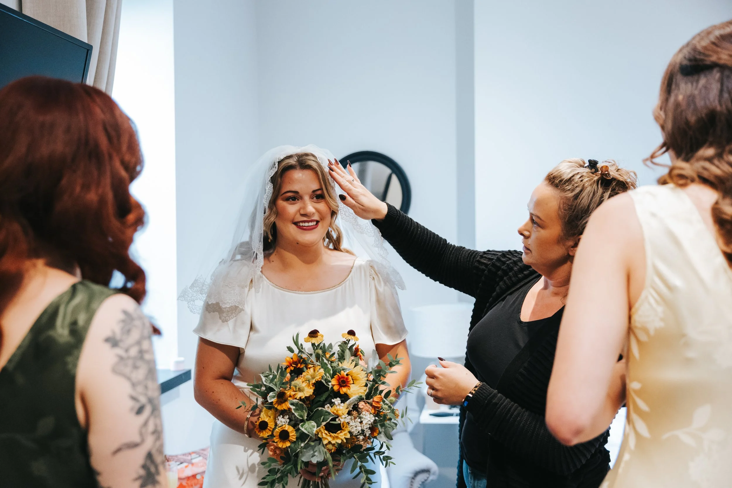 Bride with a bouquet of sunflowers and a lace veil smiling as she is assisted by a woman in black in a light-colored room with three other women nearby.