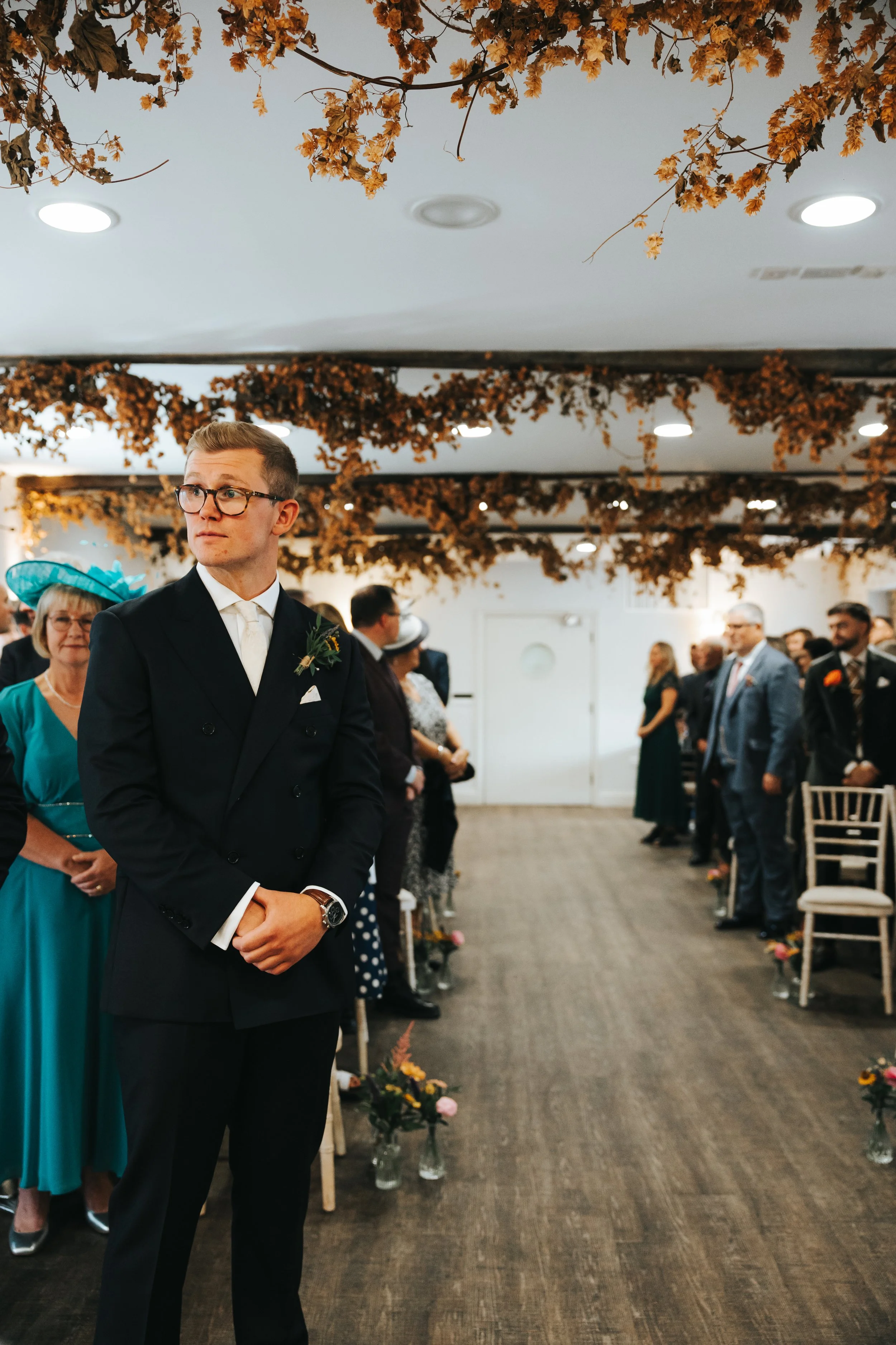 A young man in a black tuxedo with glasses and a boutonniere stands in a decorated indoor wedding venue, surrounded by seated guests, with autumn-colored floral decorations hanging from the ceiling.