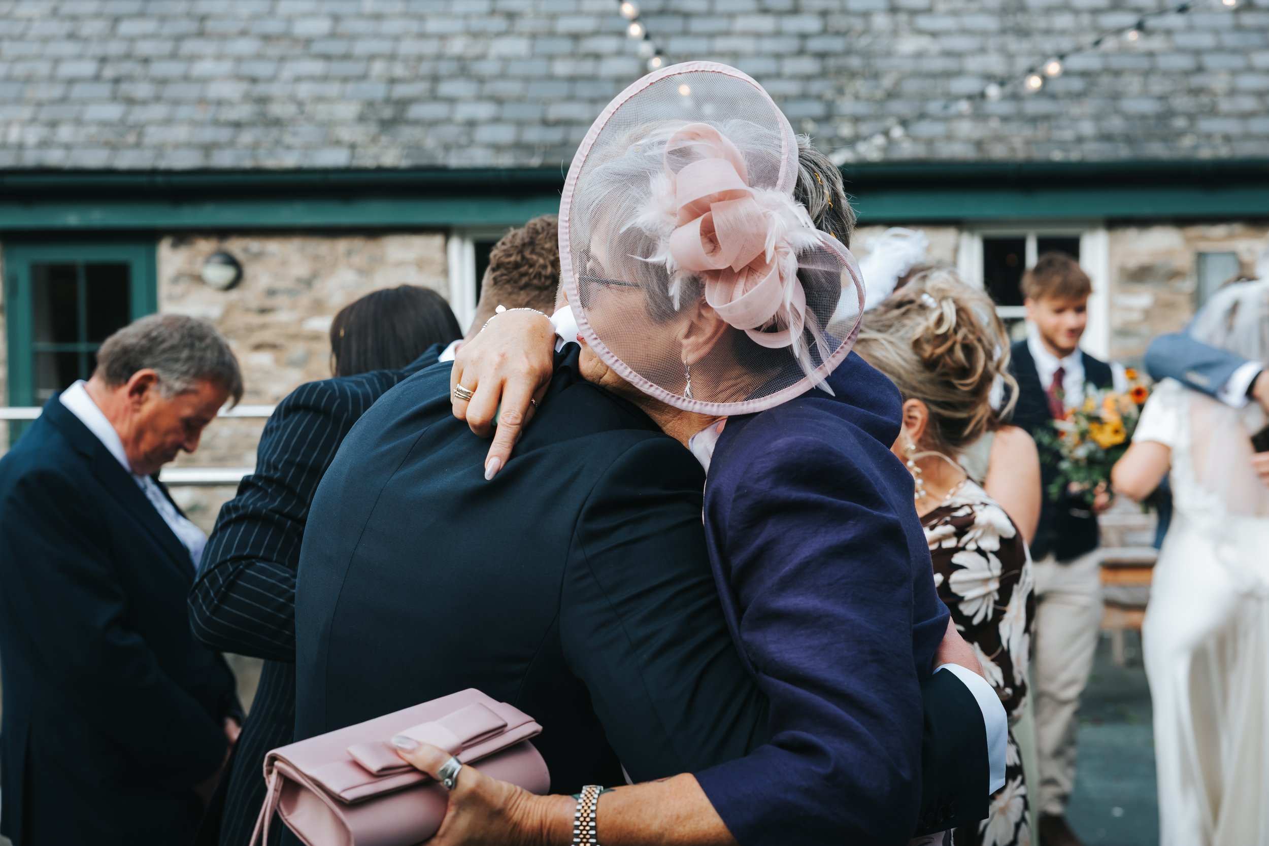 Two women hugging at a wedding, one wearing a pink fascinator and glasses, the other wearing a dark blue blazer, with other guests and a stone building in the background.