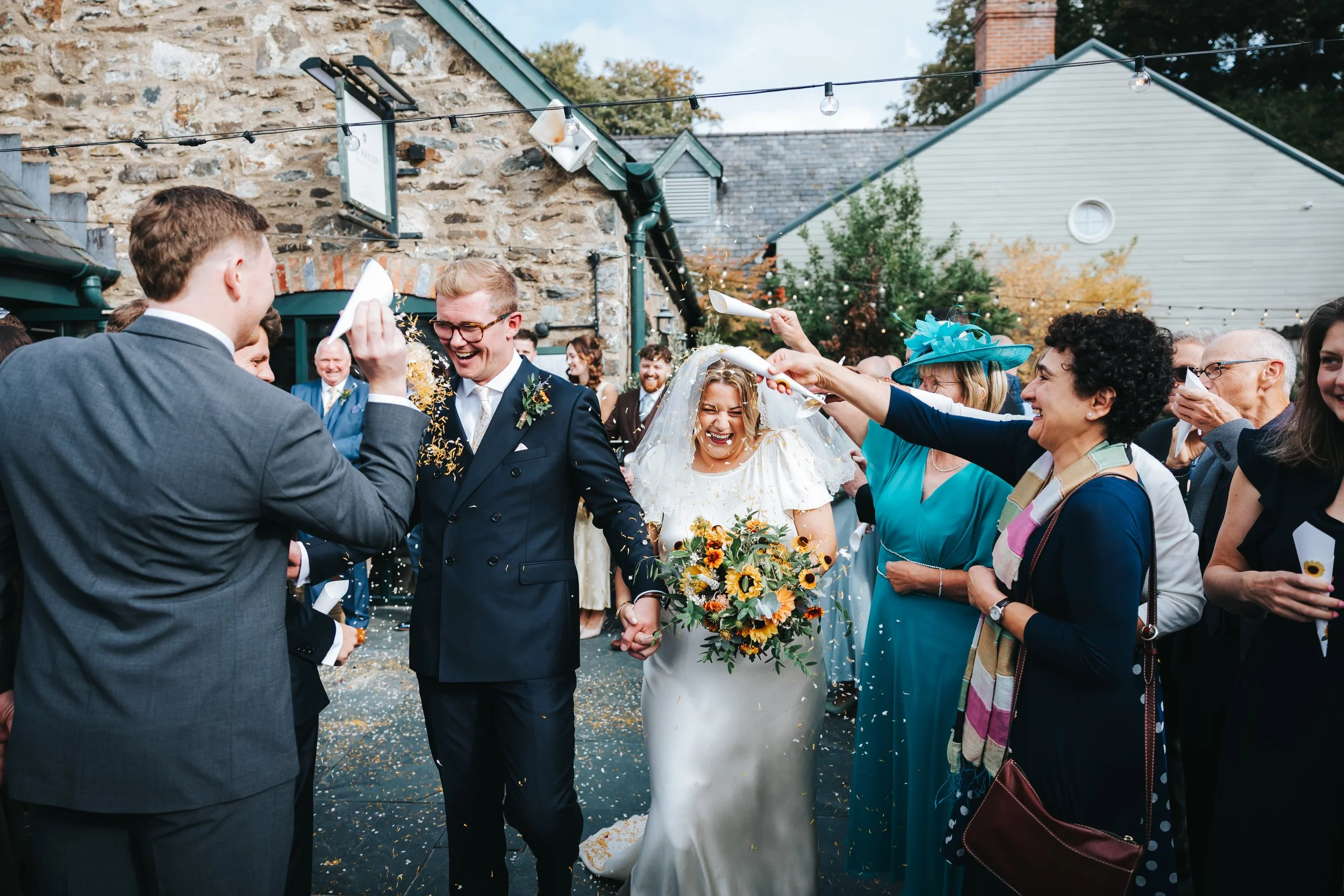 Bride and groom celebrating with guests at an outdoor wedding, with guests throwing confetti and smiling. The bride is holding a bouquet of sunflowers.