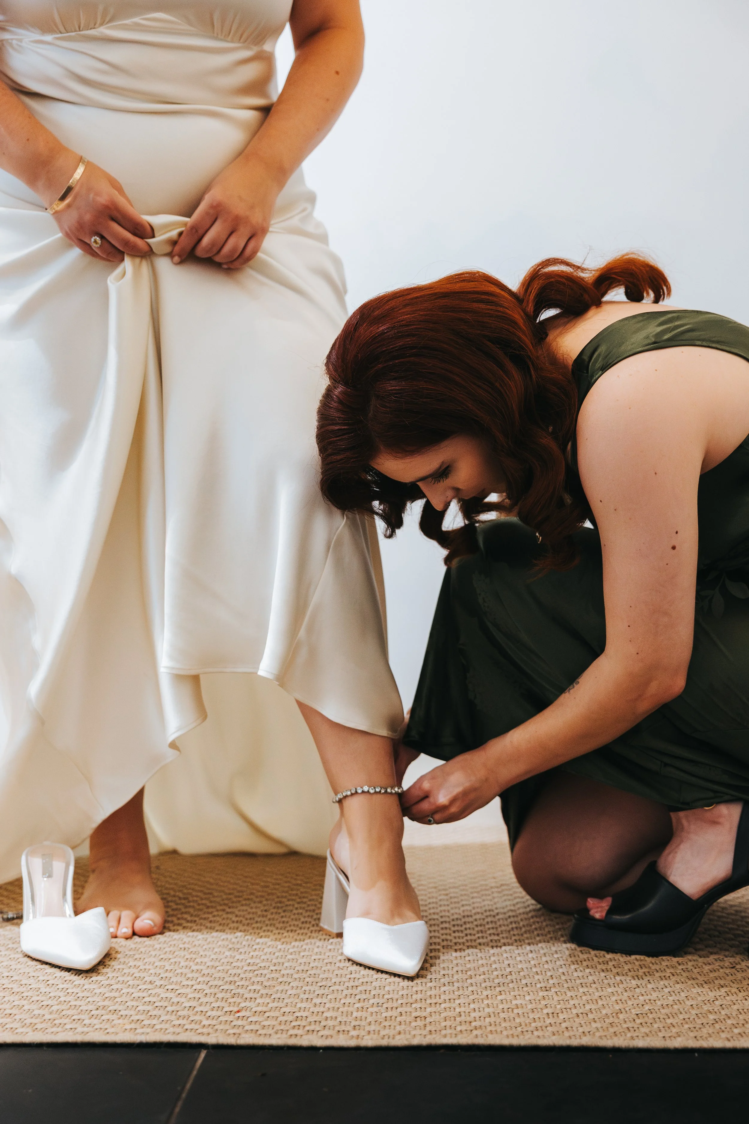 A woman in a cream-colored dress is being helped by another woman in a dark green dress to put on white high-heeled shoes.