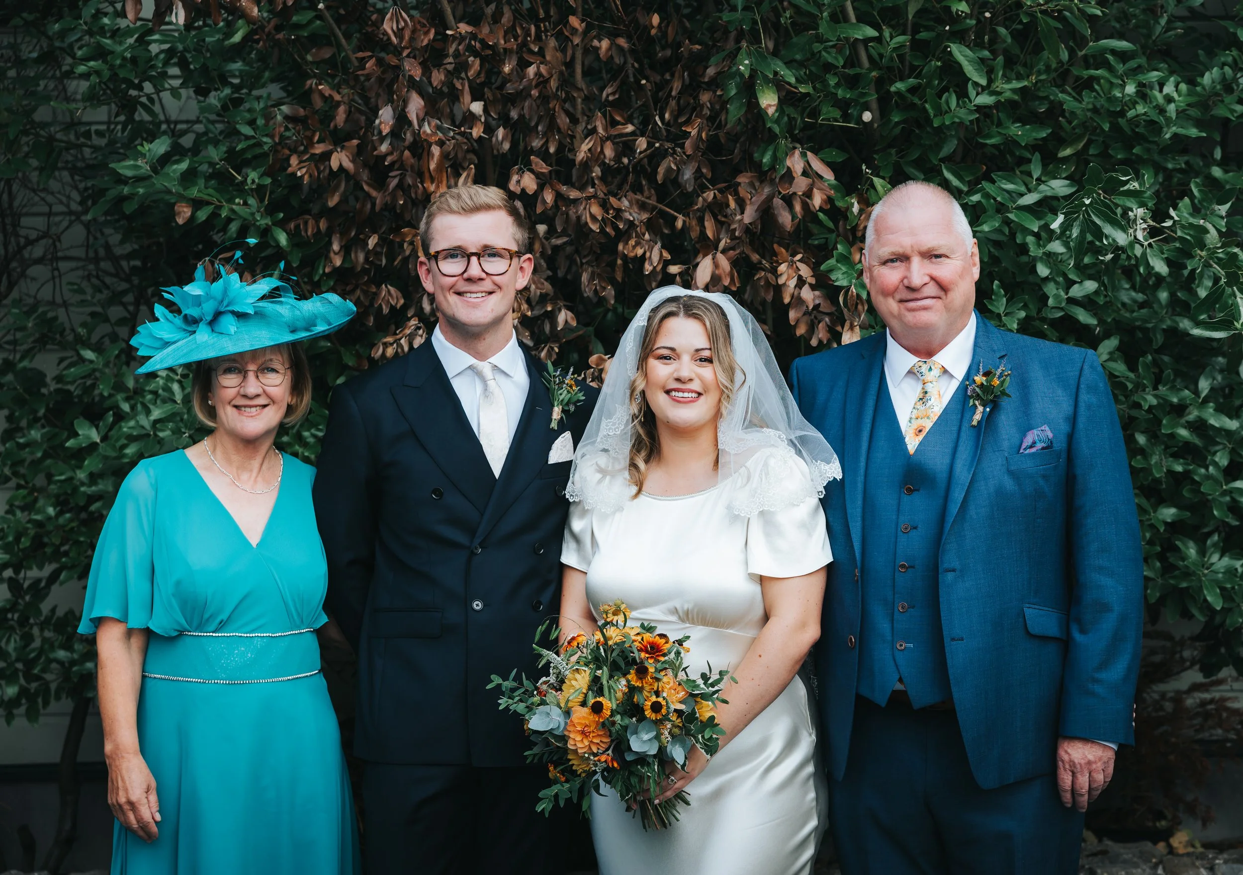 Group photo at a wedding with five people standing in front of a bush. From left to right: an older woman in a bright blue dress with a matching wide-brimmed hat, a young man in a dark suit with glasses, a smiling bride in a white dress with lace det