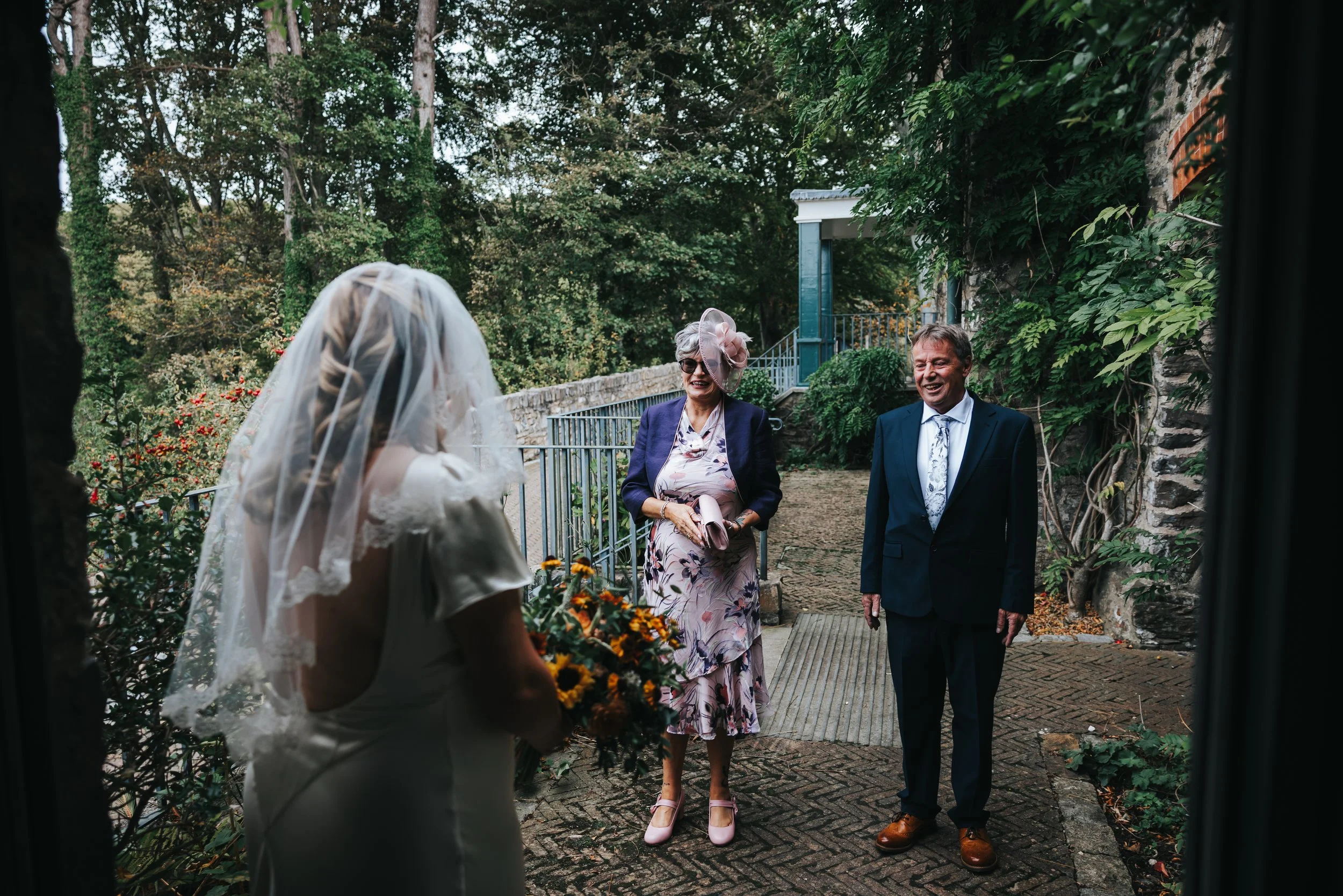 A bride facing her parents in an outdoor garden setting with trees, bushes, and a stone building, during a wedding ceremony or greeting.
