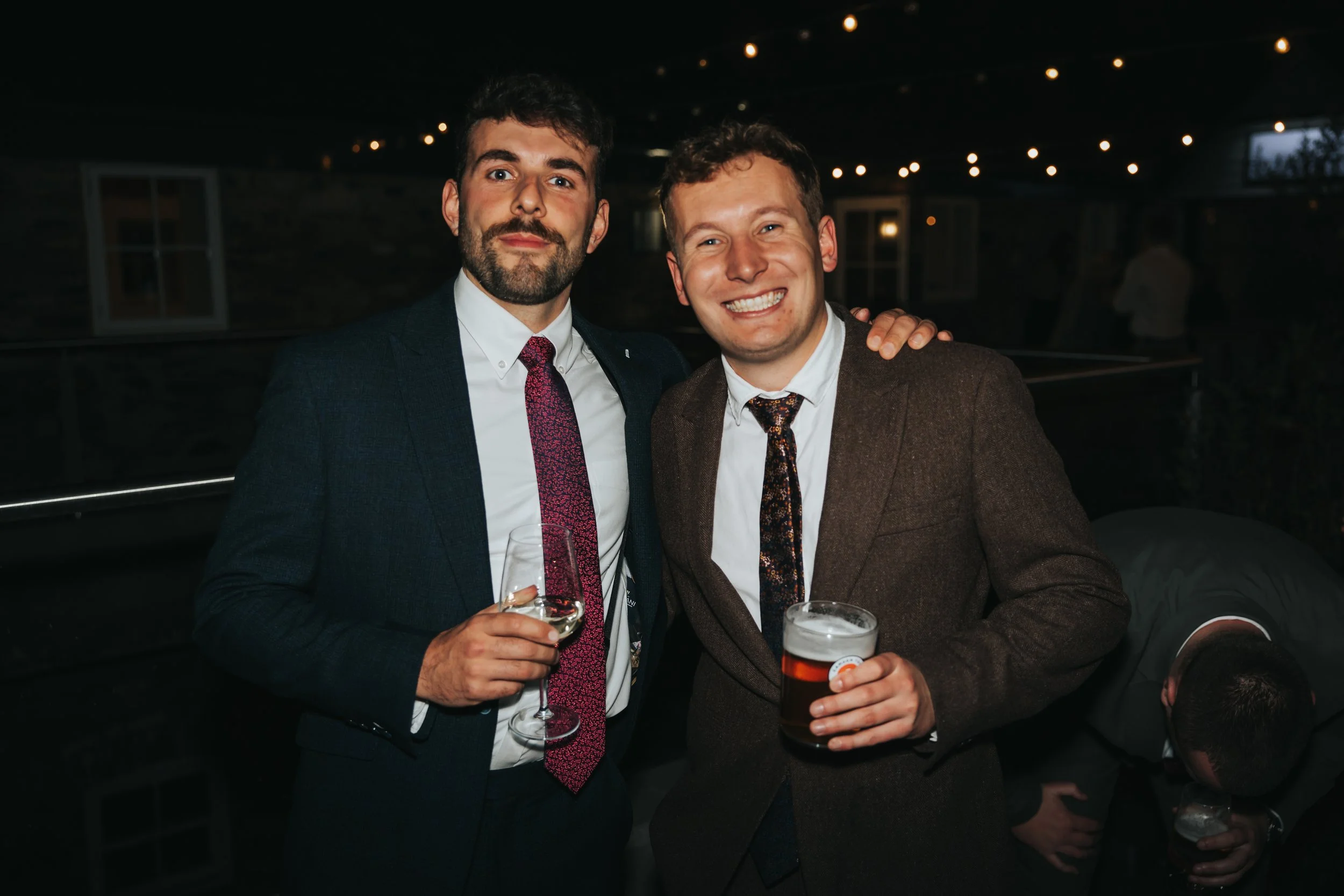 Two men in suits holding drinks, smiling, at a social event with dim lighting and string lights overhead.