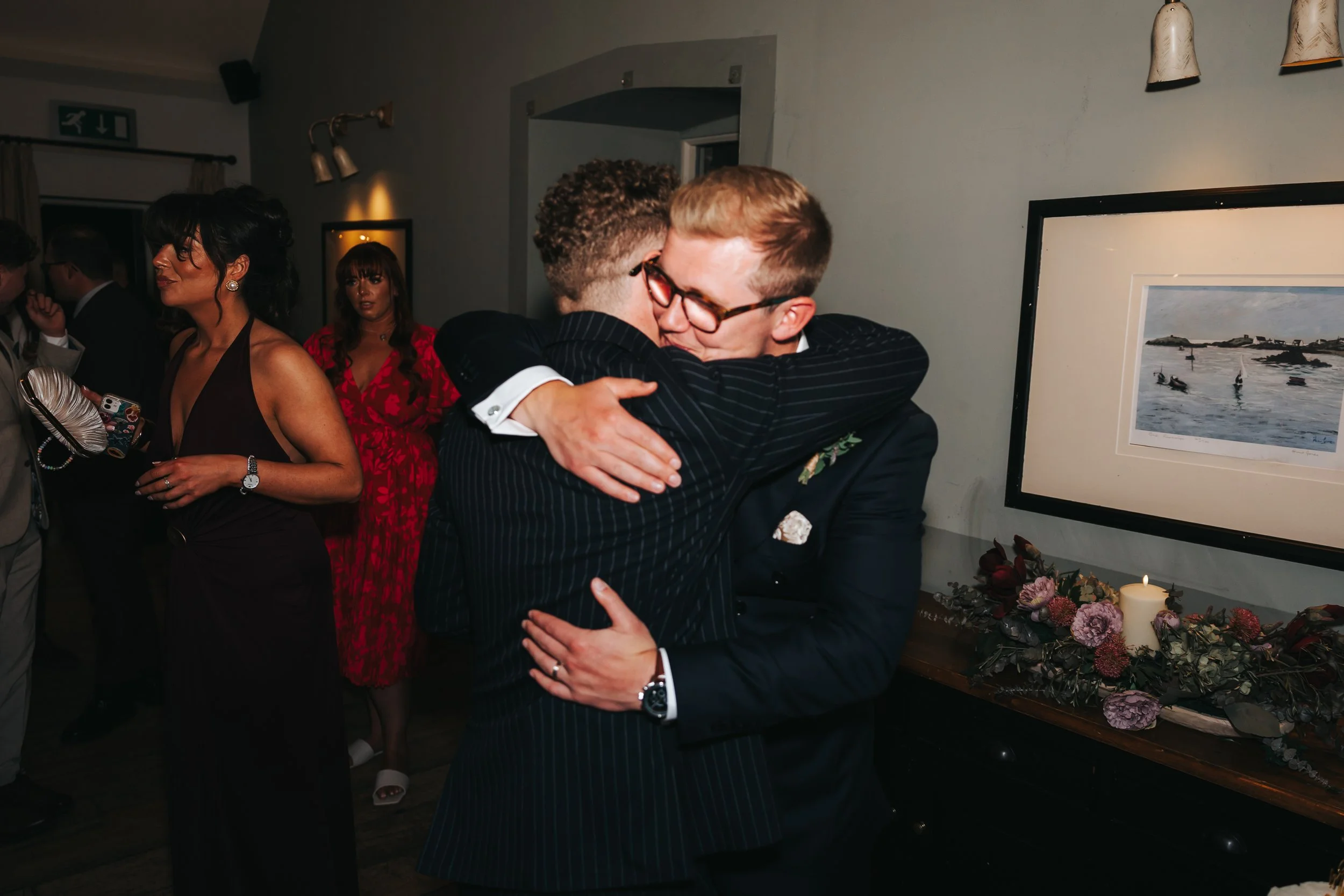 Two men hugging at a social event, with other people in the background, including a woman in a red dress and a woman in a black dress holding a clutch, in a decorated indoor space.