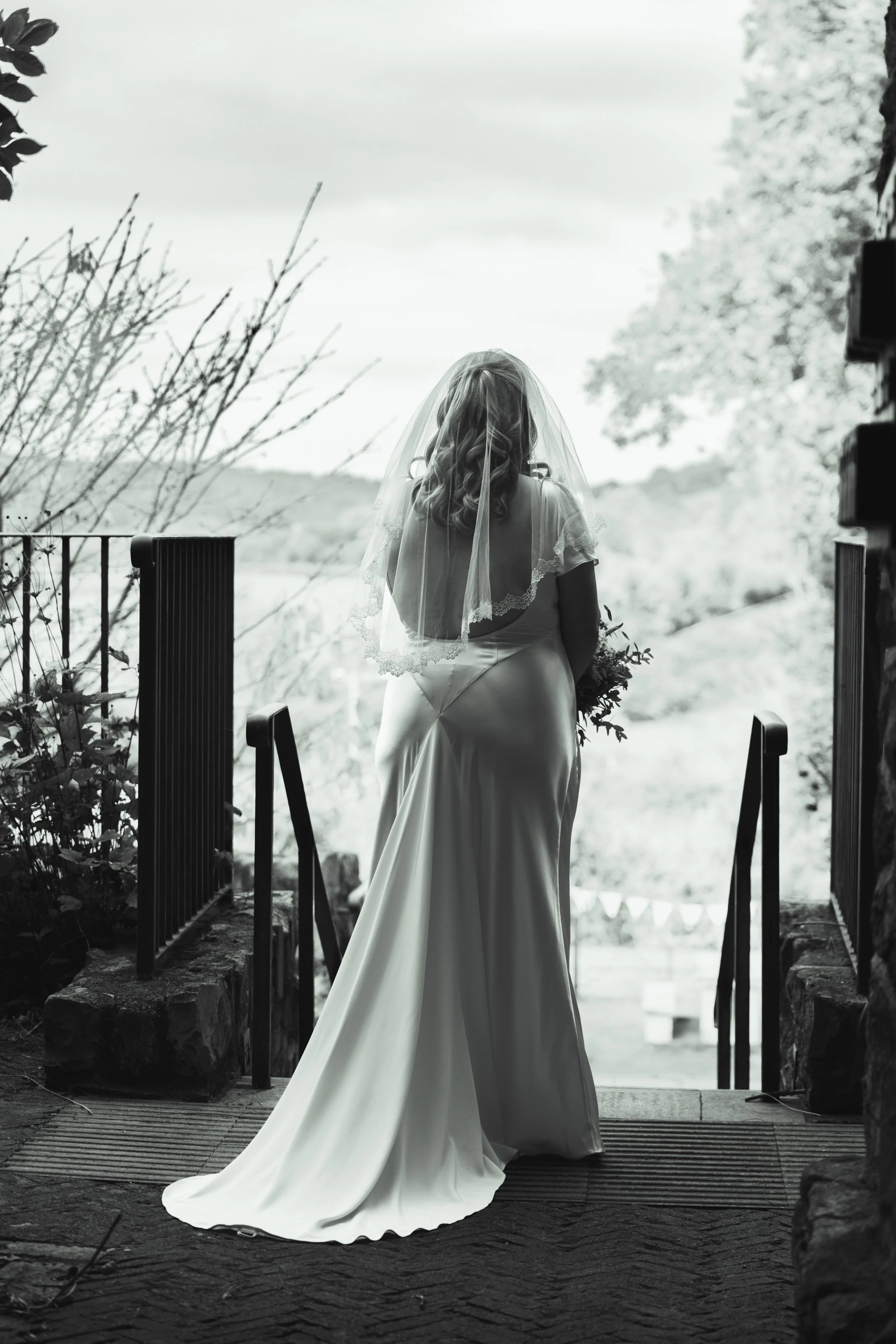 Black-and-white photo of a bride in a wedding gown and veil looking out from a porch or balcony.