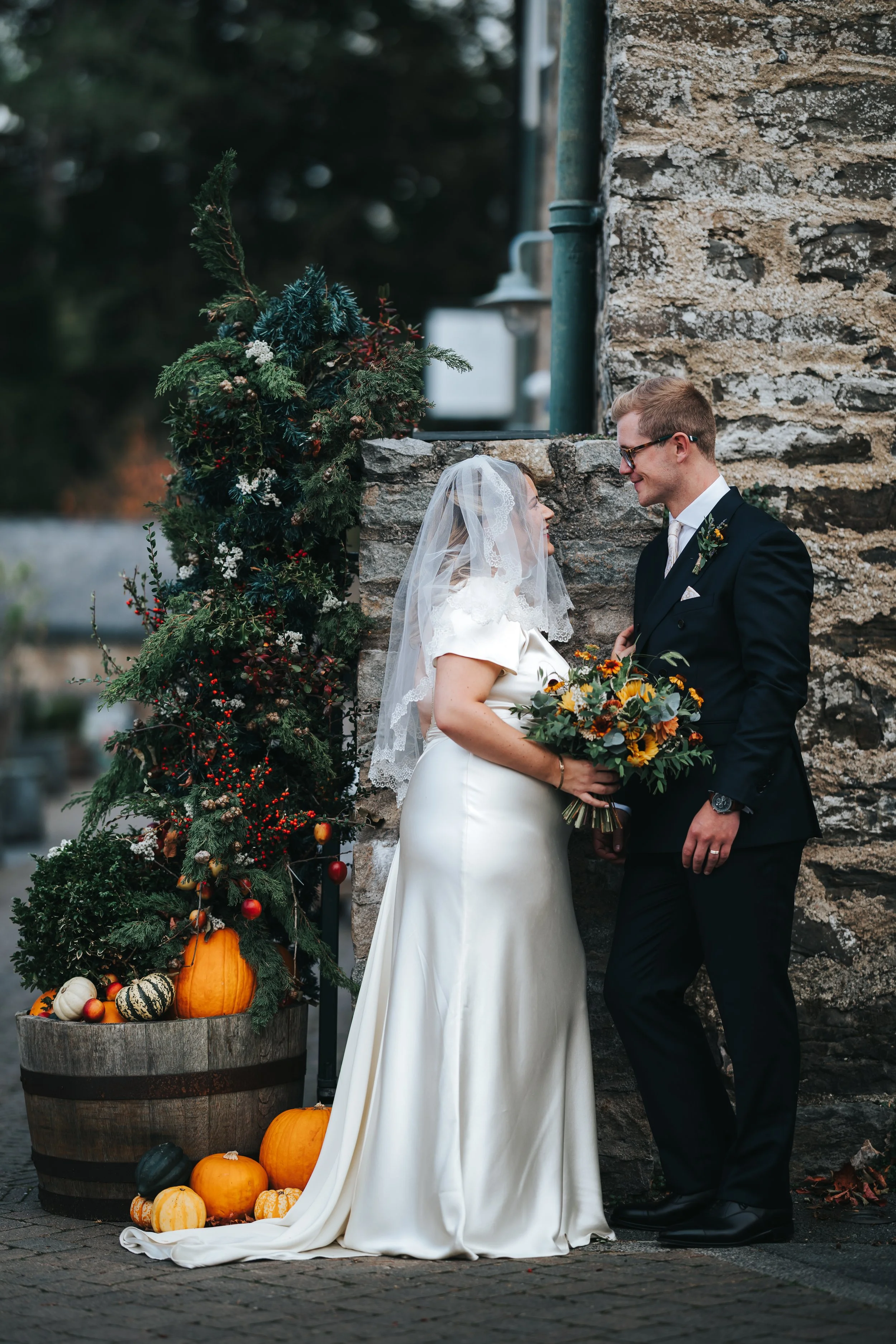 A bride and groom standing close together outdoors, with a decorated fall-themed pumpkin and foliage arrangement beside them.