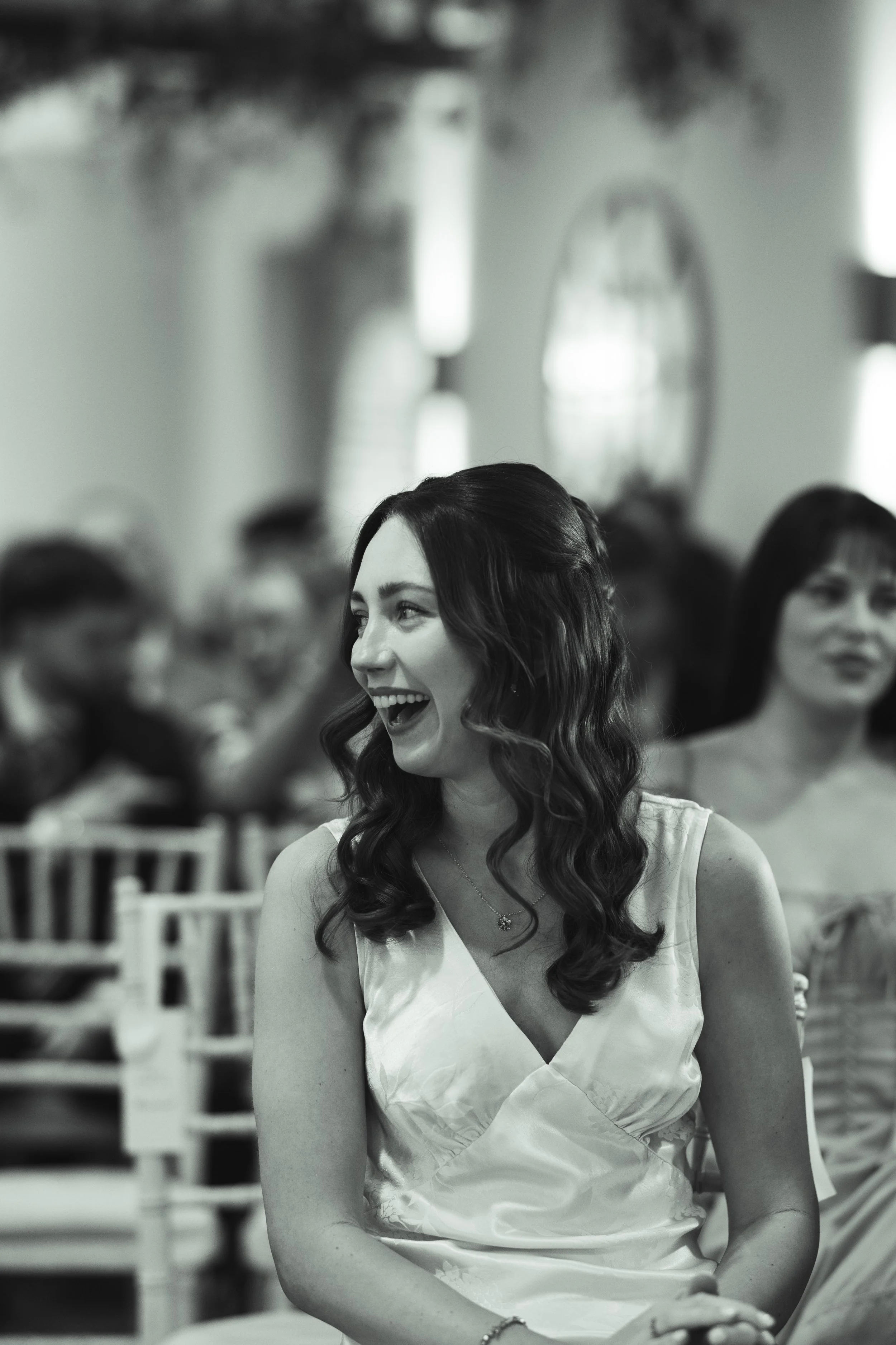 A woman with long dark wavy hair, smiling and looking to the side, seated at an indoor event with blurred people in the background.