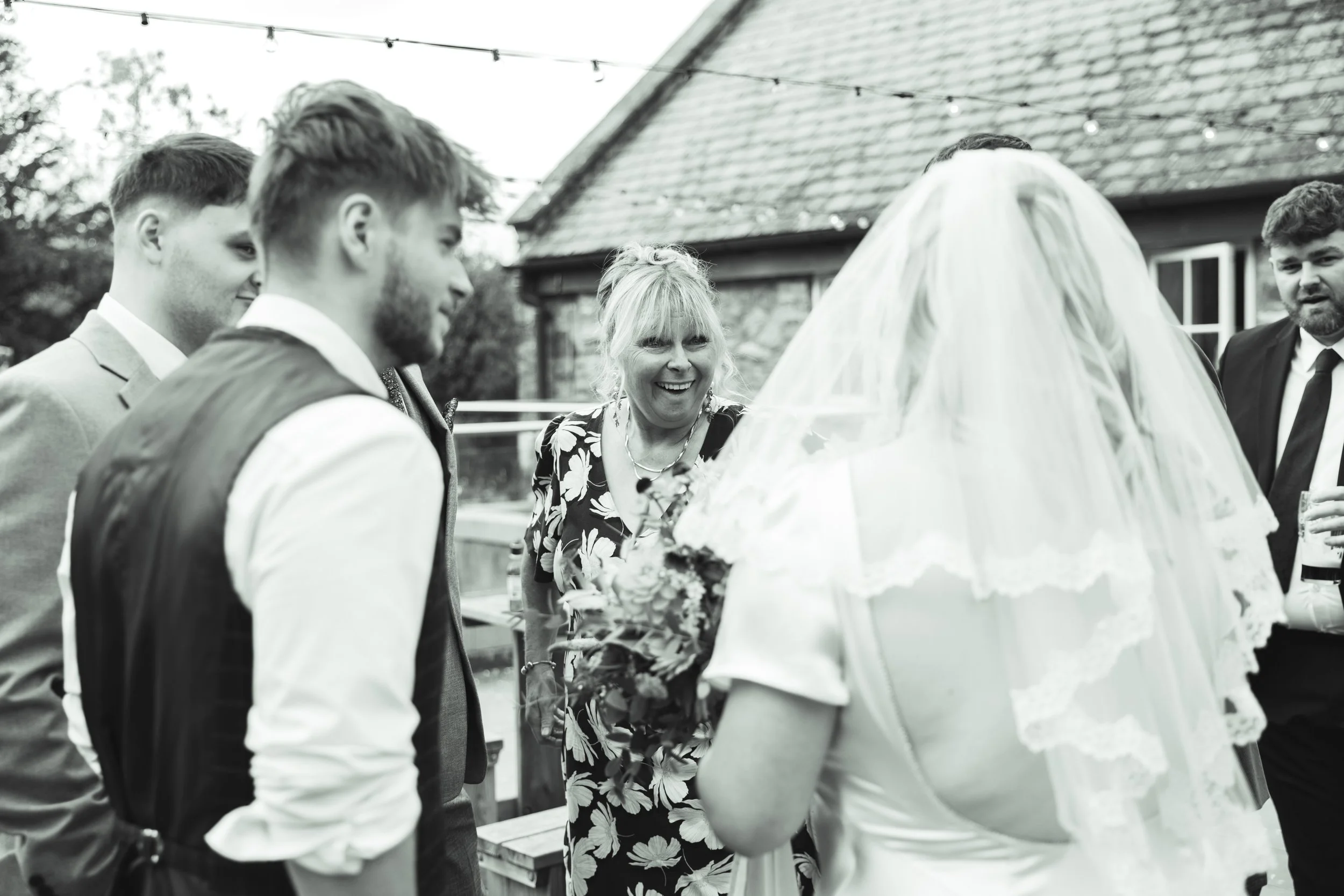 A bride with a lace veil is greeting a group of men in suits and a woman in a patterned dress at an outdoor wedding celebration.