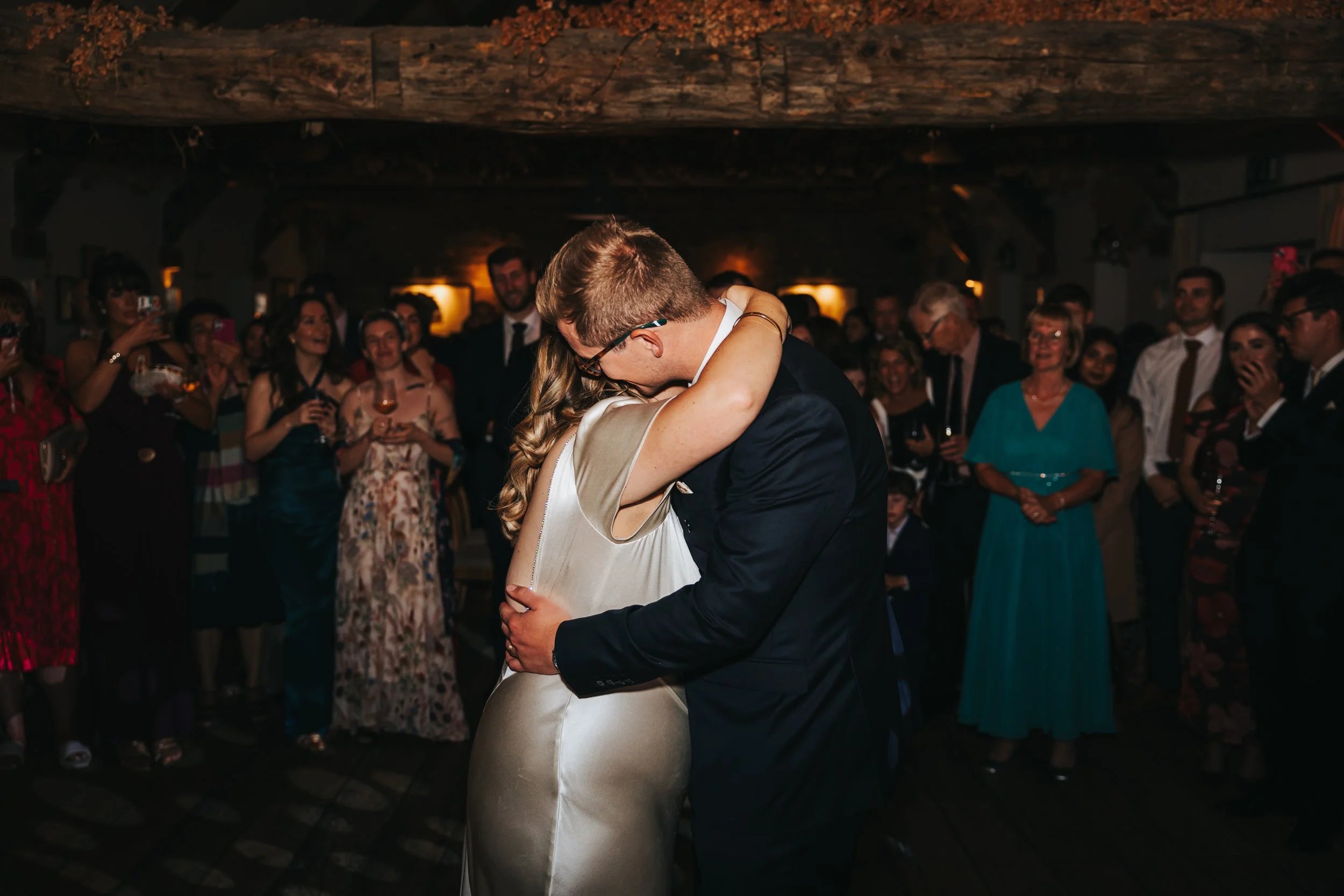 A newlywed couple shares their first dance at the wedding reception in a dimly lit rustic venue, surrounded by guests watching and capturing the moment.