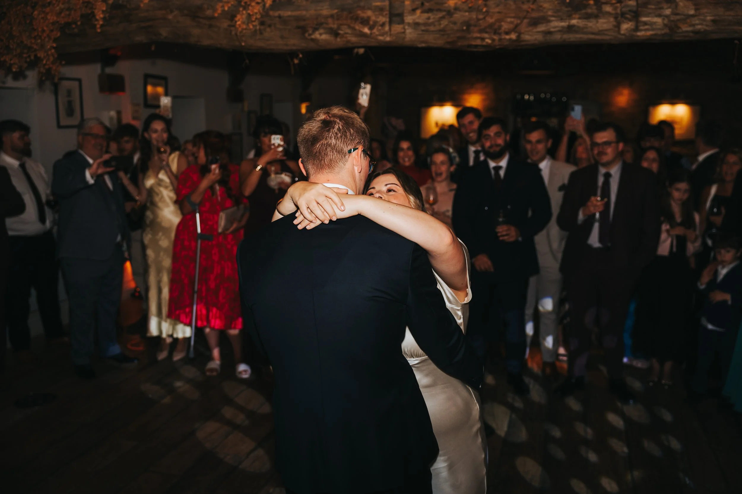 Couple dancing closely during a wedding reception surrounded by guests watching and taking photos.