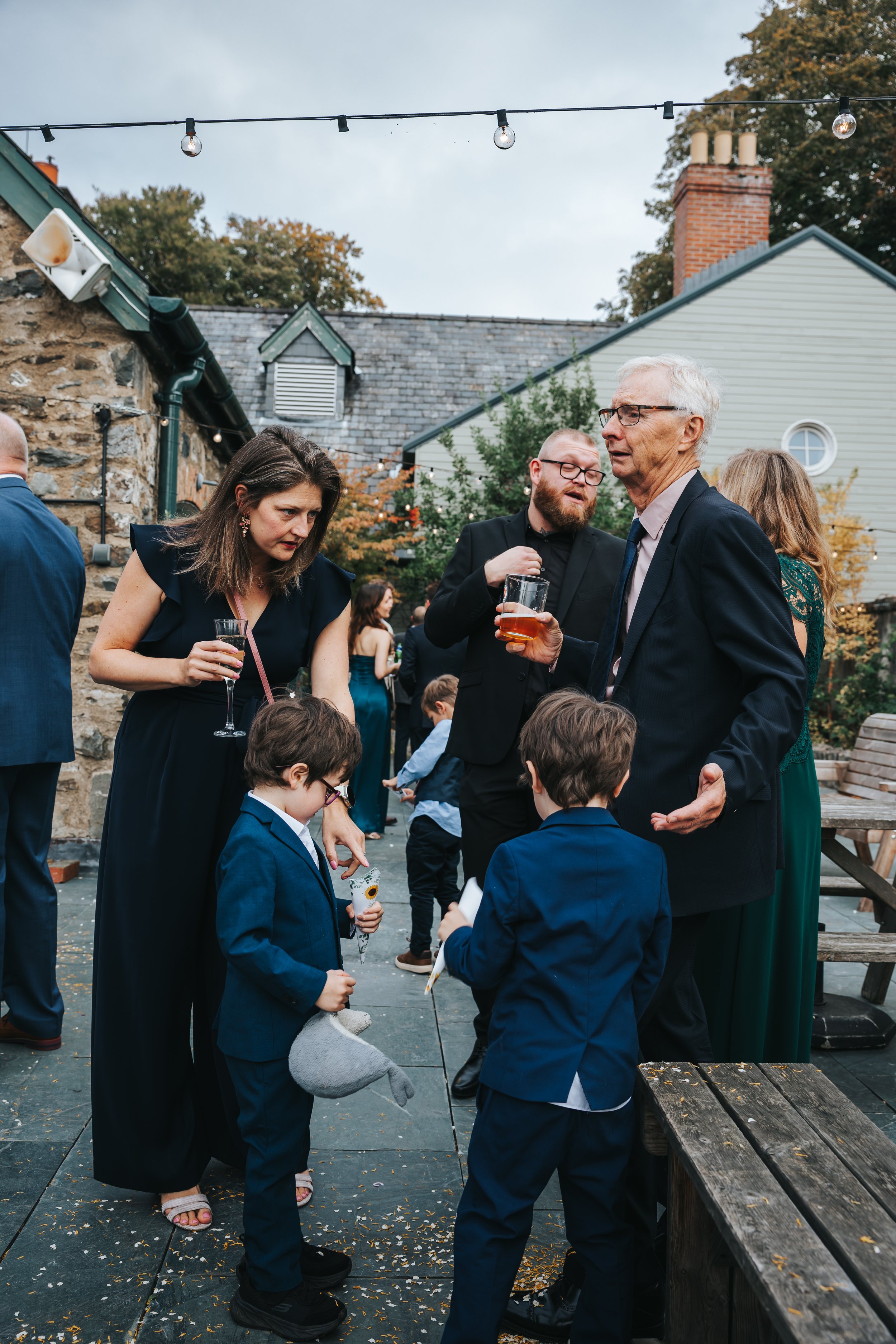 People gathered outdoors at a social event or party, talking and holding drinks, with children present, during the evening with string lights overhead.