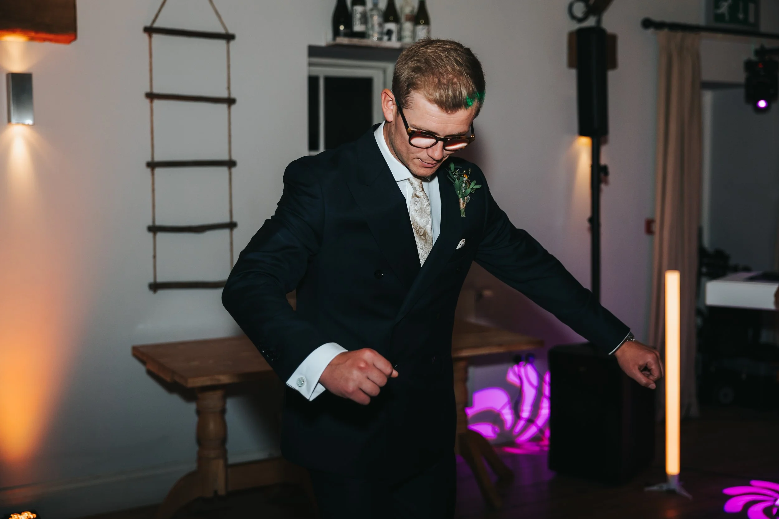 Man in a dark suit and glasses dancing at an indoor event with colorful lighting.