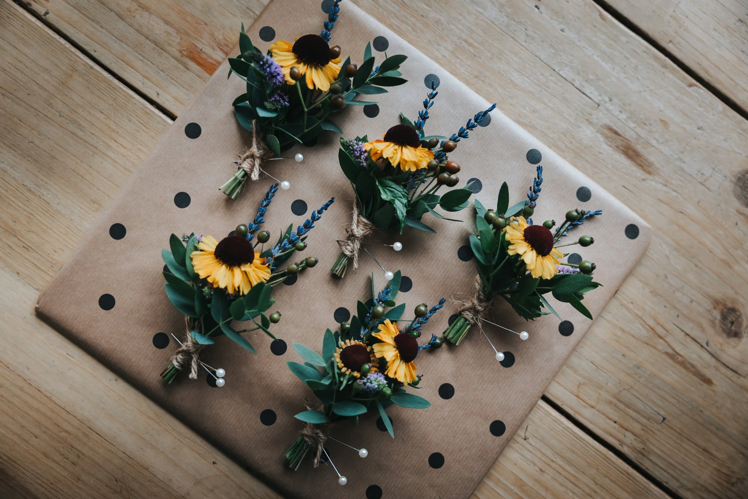 Five small floral bouquets with yellow flowers, blue lavender, green leaves, and small white pearl accents arranged on a wrapped gift with black polka dots on kraft paper, placed on a wooden surface.