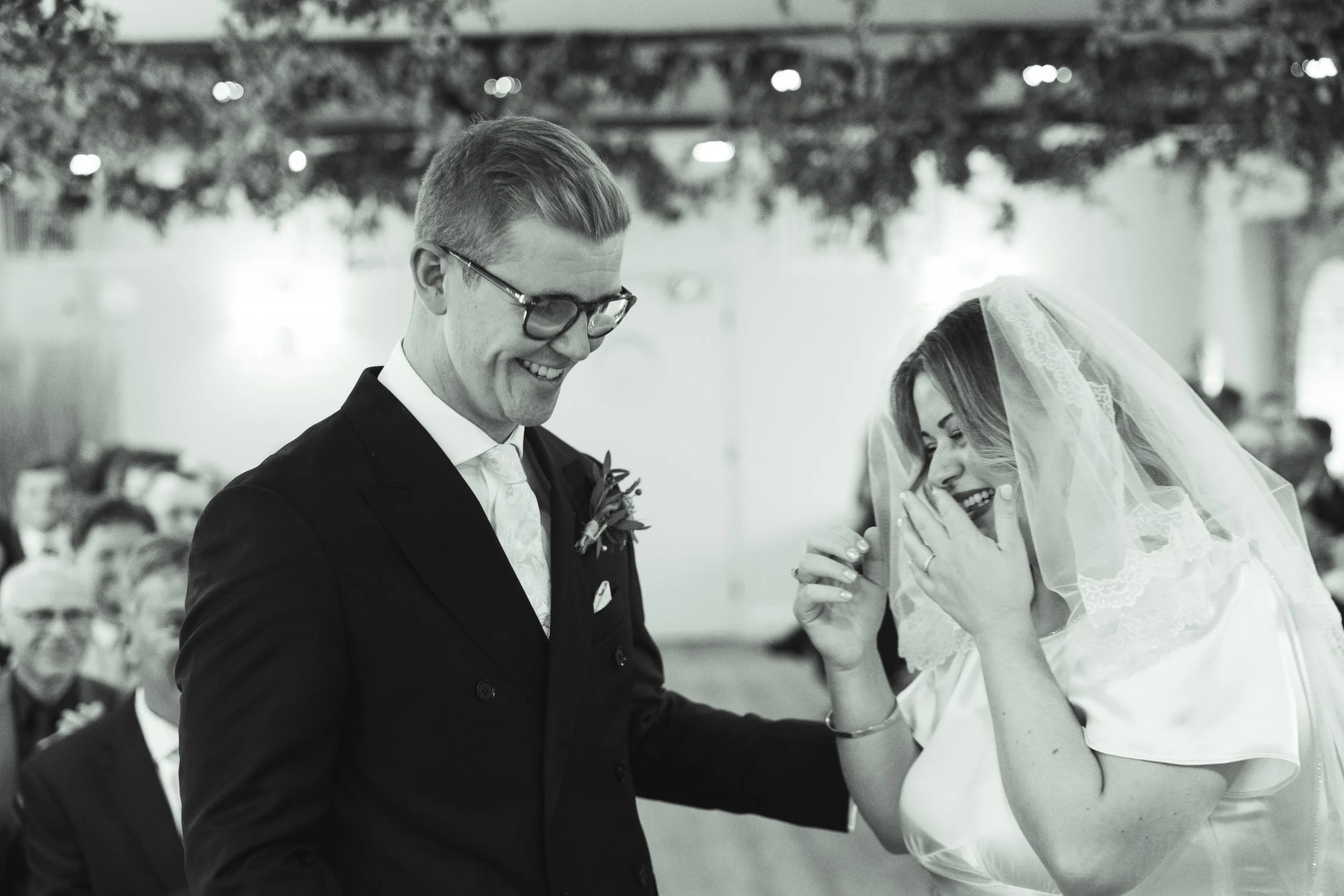 A black-and-white photo of a wedding ceremony showing a groom in a tuxedo with glasses smiling, and a bride with a lace veil covering her face, covering her mouth with her hand in a moment of joy.