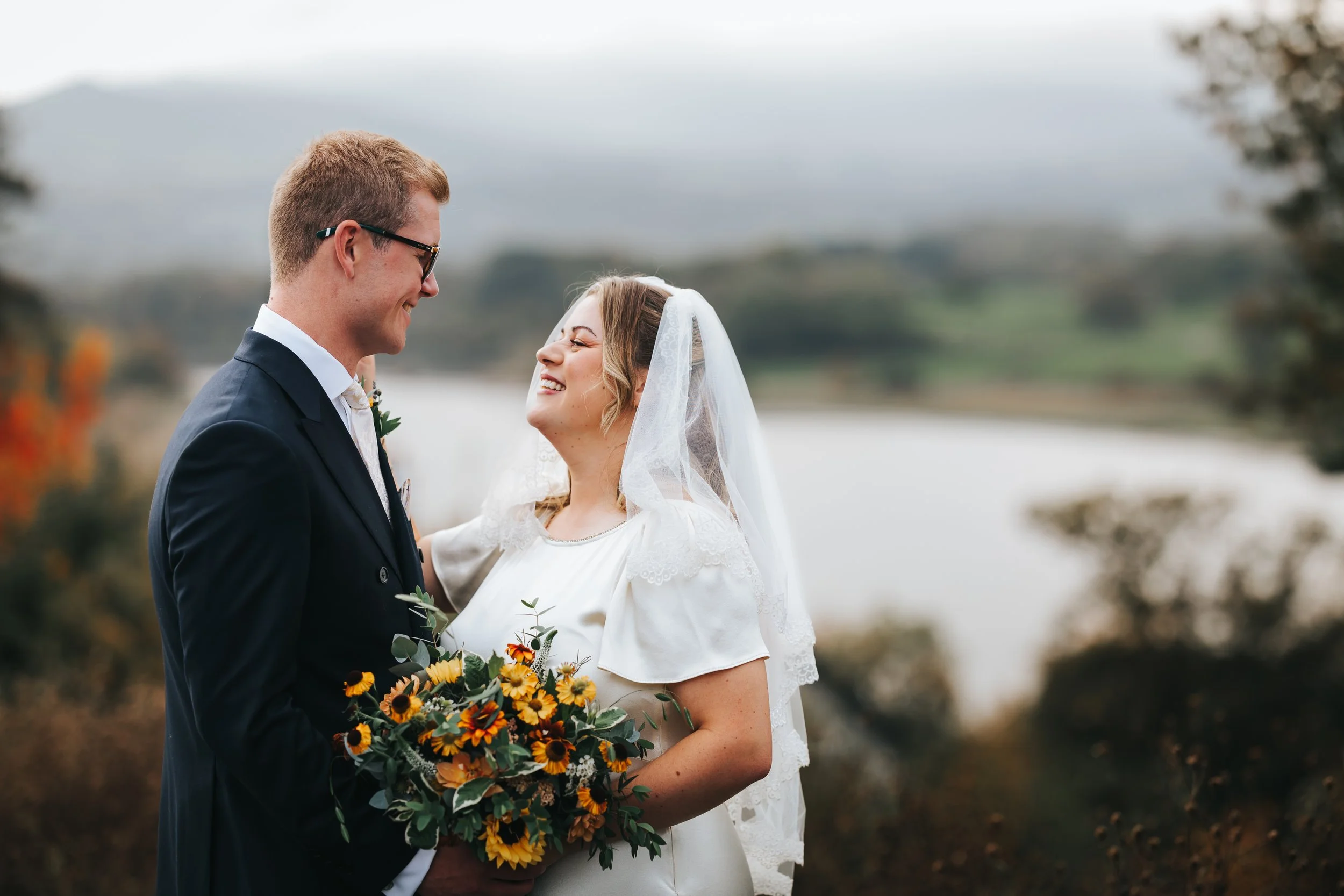 A bride and groom sharing a moment outdoors with a lake and hills in the background, the bride holding a bouquet of sunflowers and wearing a veil, and the groom dressed in a dark suit with sunglasses.