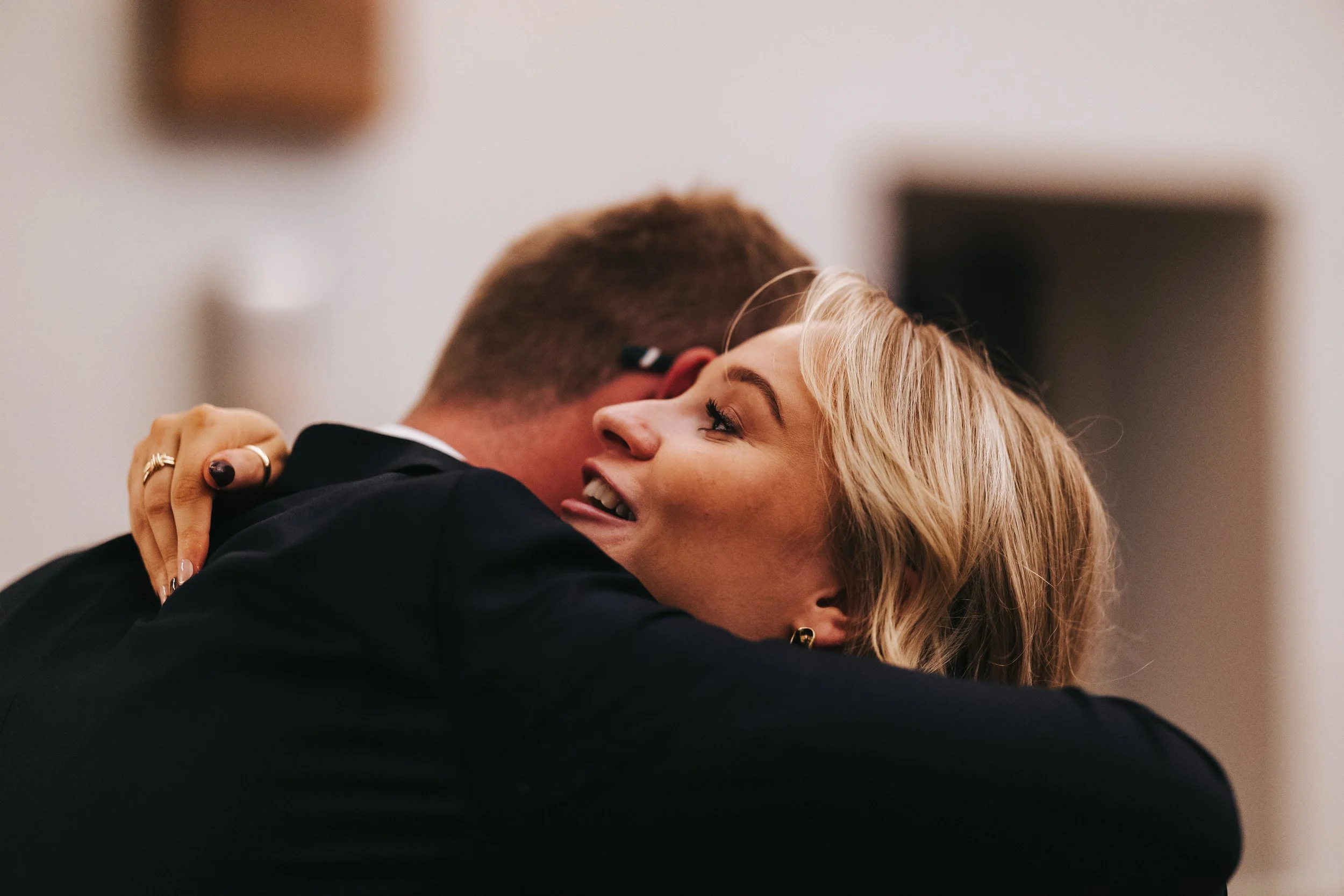 A woman with blonde hair is hugging a man in a black suit, smiling and closing her eyes, in an emotional moment.