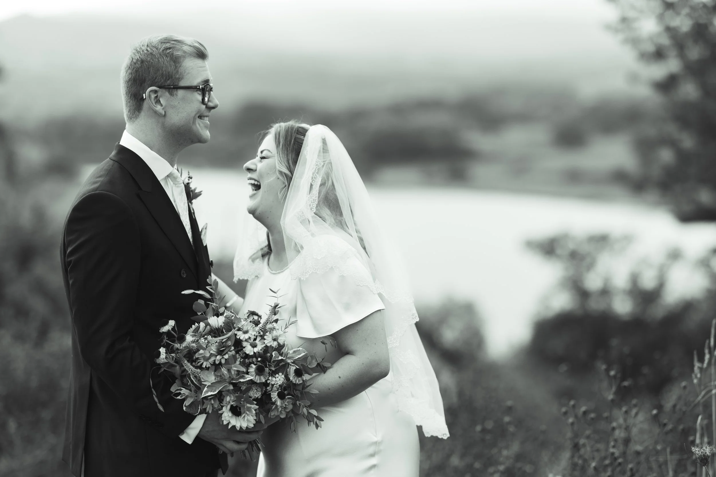 Black and white photo of a bride and groom smiling and looking at each other outdoors, with the bride holding a bouquet of flowers.