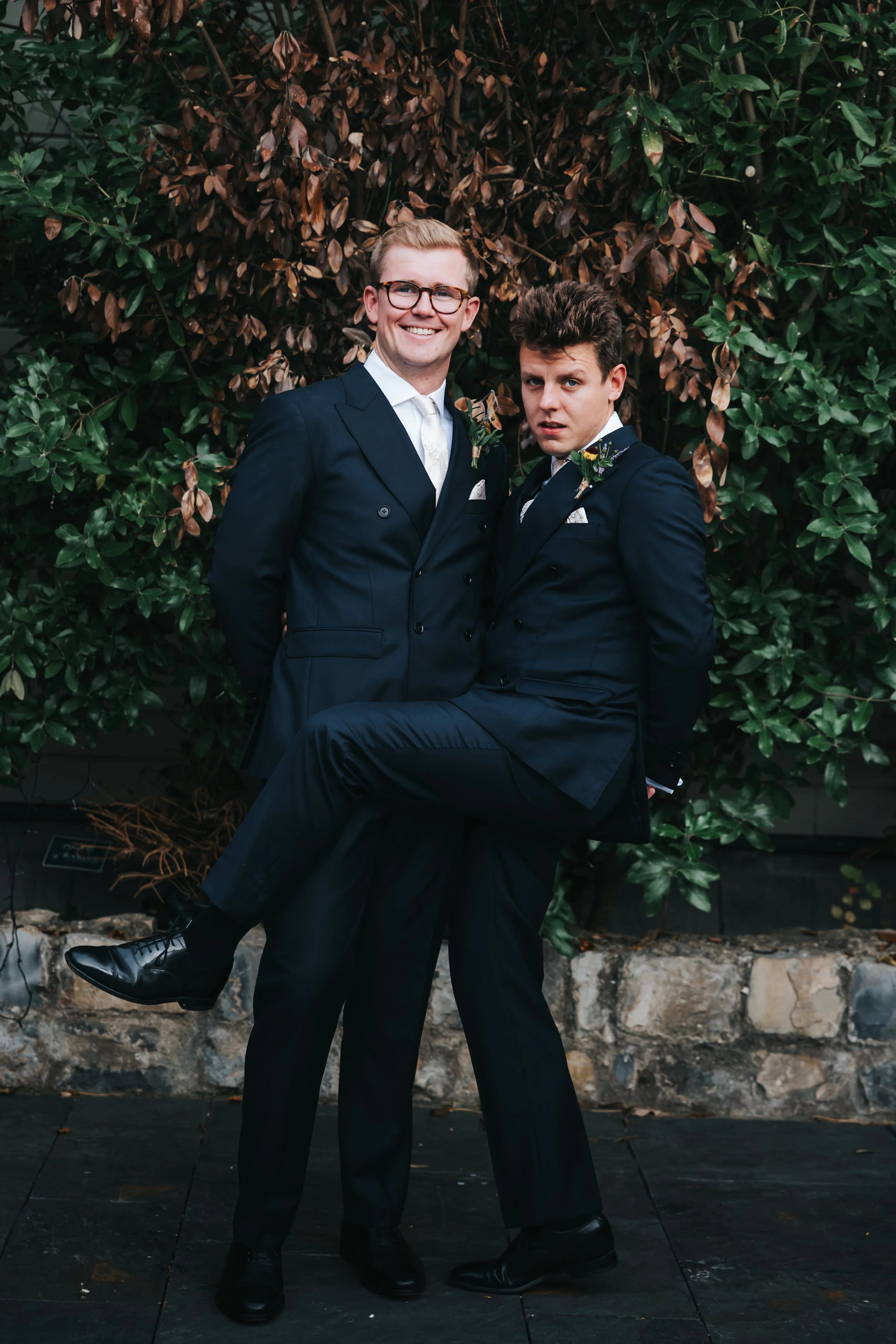 Two men in formal suits and ties posing outdoors in front of a leafy green and brown shrub, one lifting the other.
