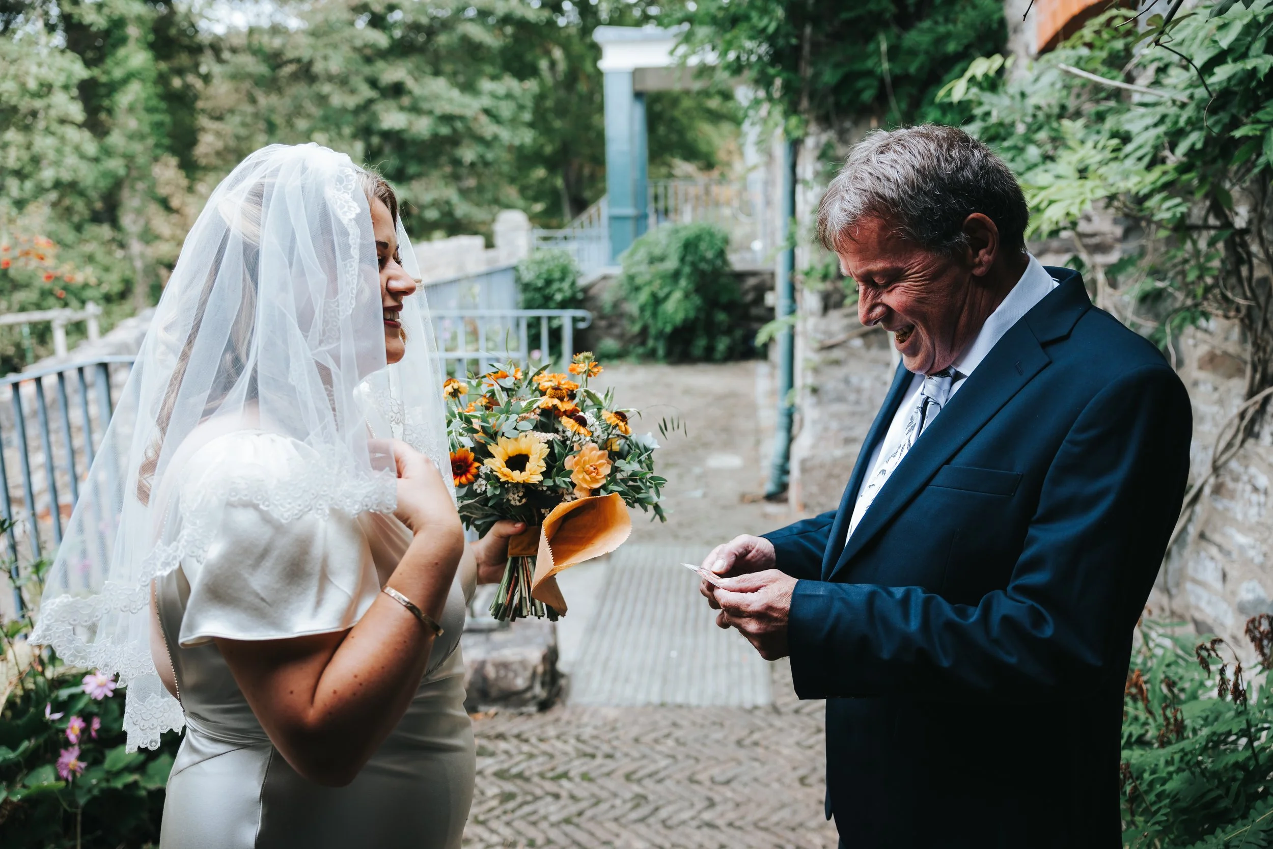 A bride in a white dress and veil holding a bouquet of flowers stands outdoors next to an older man in a suit who is smiling as he looks at a piece of paper, possibly during a wedding ceremony or vow exchange.