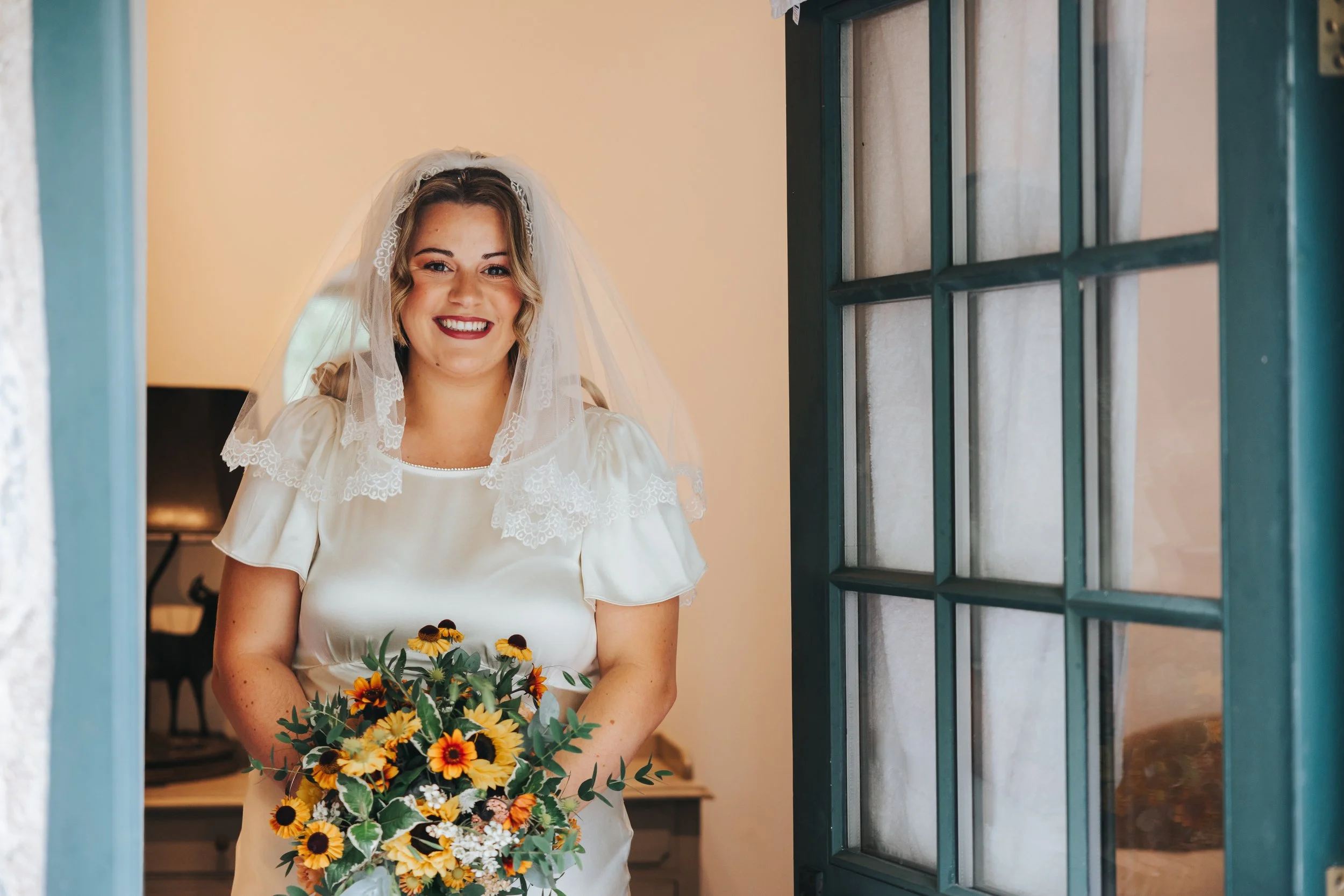 A smiling bride in a white satin dress holding a bouquet of yellow and orange flowers, standing indoors near a window.