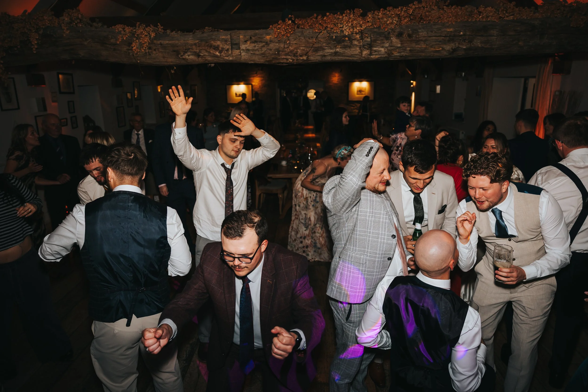 People dancing and socializing at a lively party or celebration indoors, with warm lighting and rustic decor.