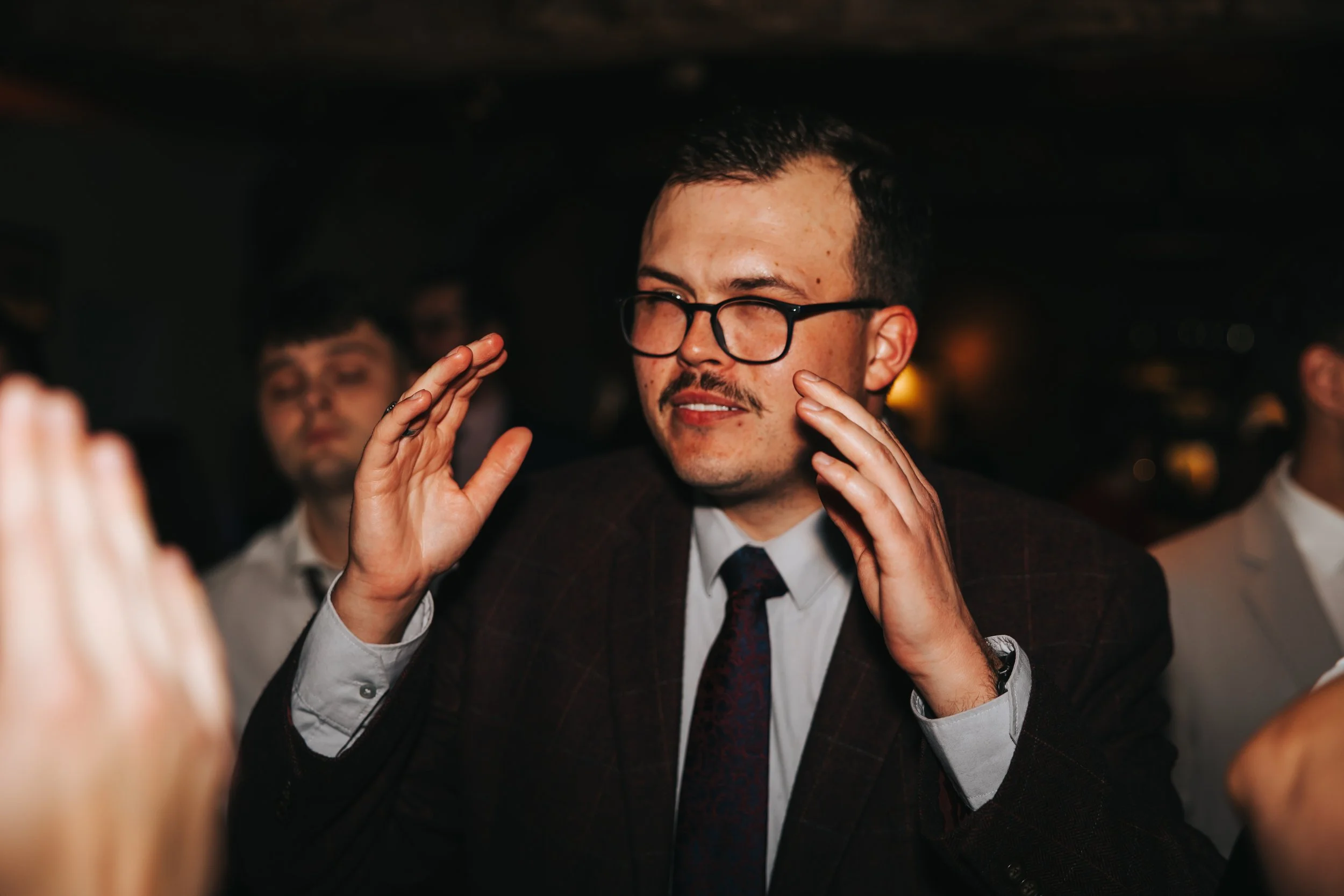 A man with glasses and a mustache, dressed in a dark checkered suit and white shirt, is touching his face with his hands in a lively setting, possibly at a social event or party.
