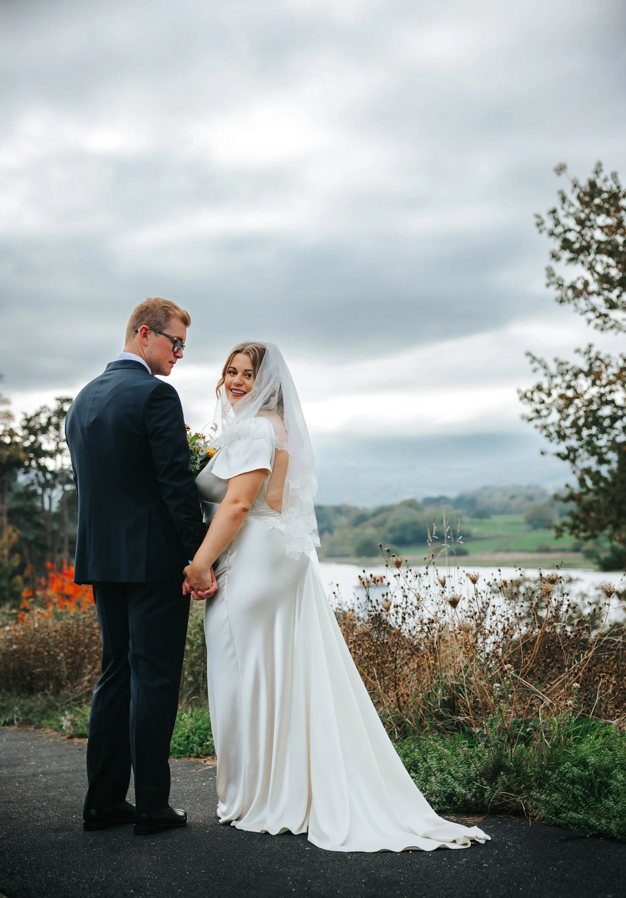 A bride and groom holding hands outdoors by a lake, with cloudy sky and trees in the background, celebrating their wedding.