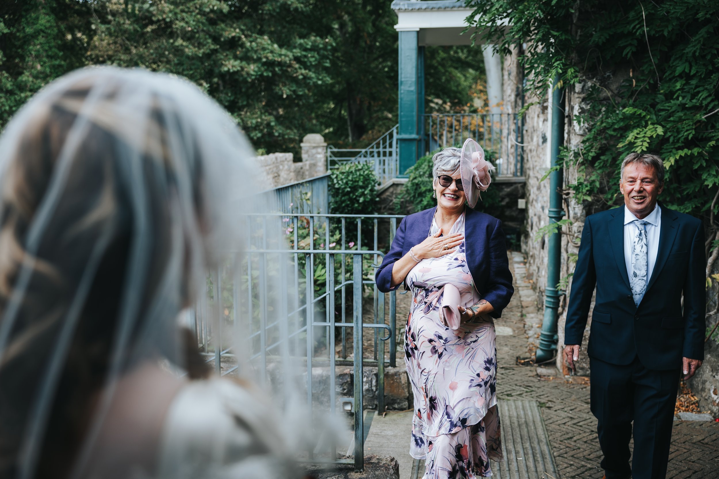 Older woman in a floral dress and pink hat happily greeting a smiling woman with long blonde hair in a wedding dress, with a man in a dark suit nearby, outdoors on a garden pathway.