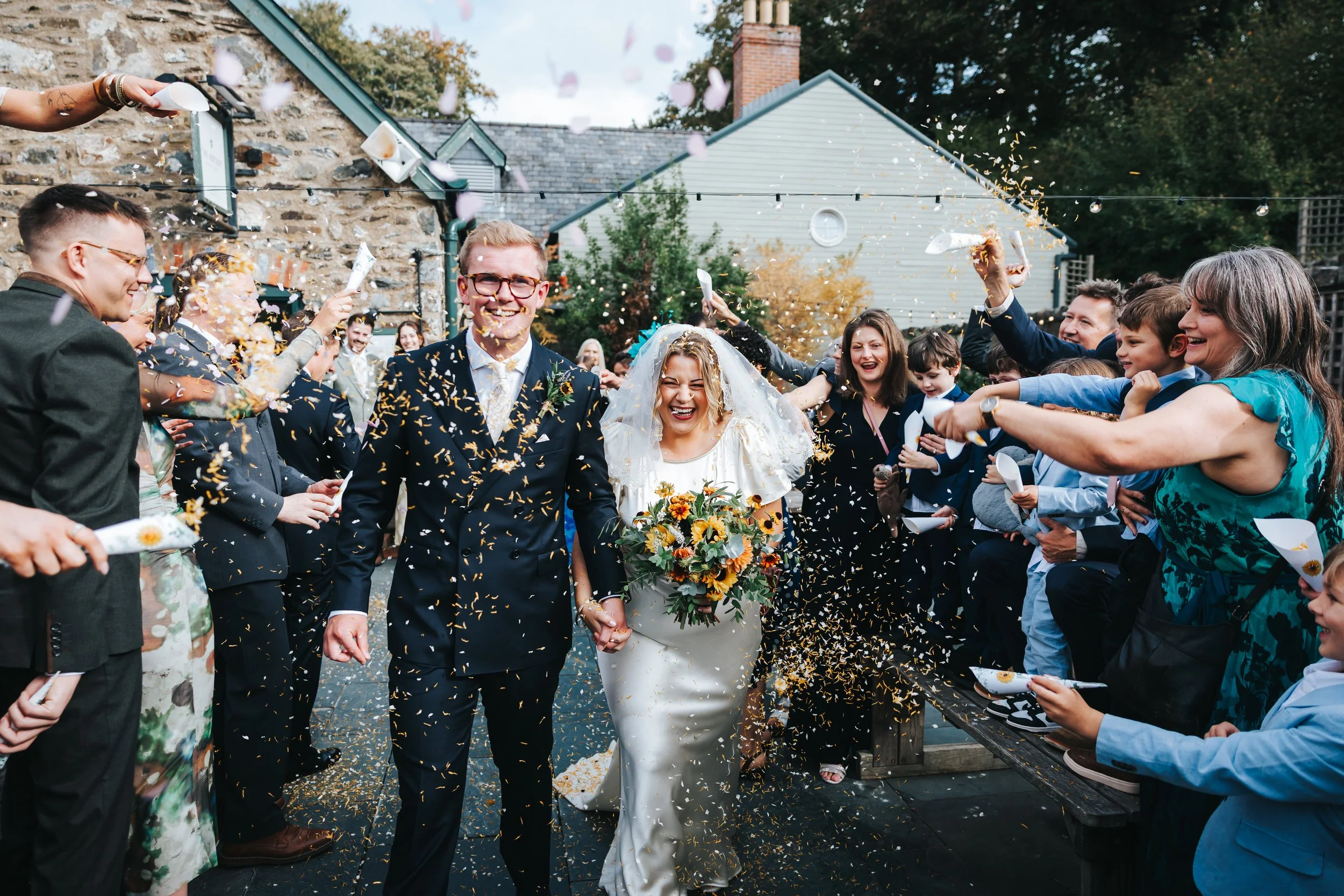 A newlywed couple walking hand in hand through a crowd throwing confetti during an outdoor wedding celebration, smiling joyfully.
