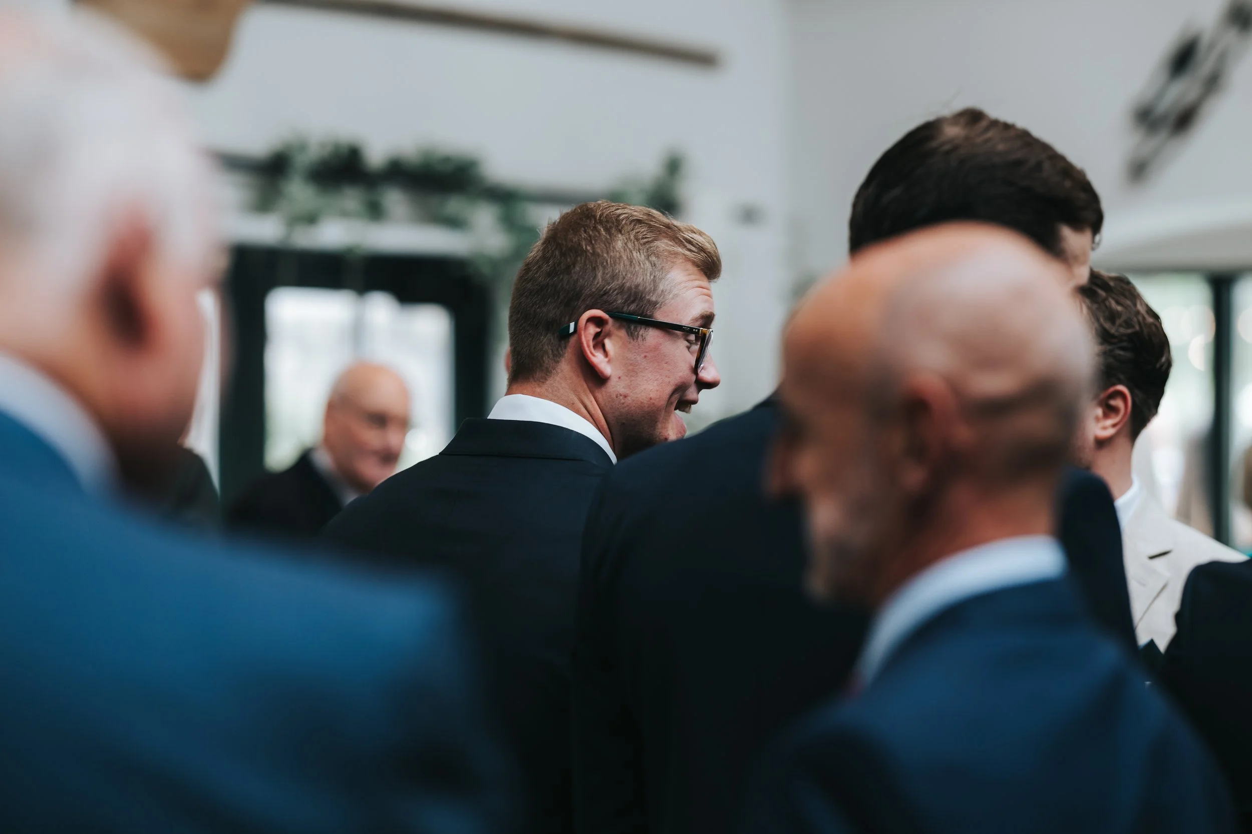 Men dressed in formal suits gathered at an indoor event, some engaged in conversation, with a focus on a man wearing glasses in the center.